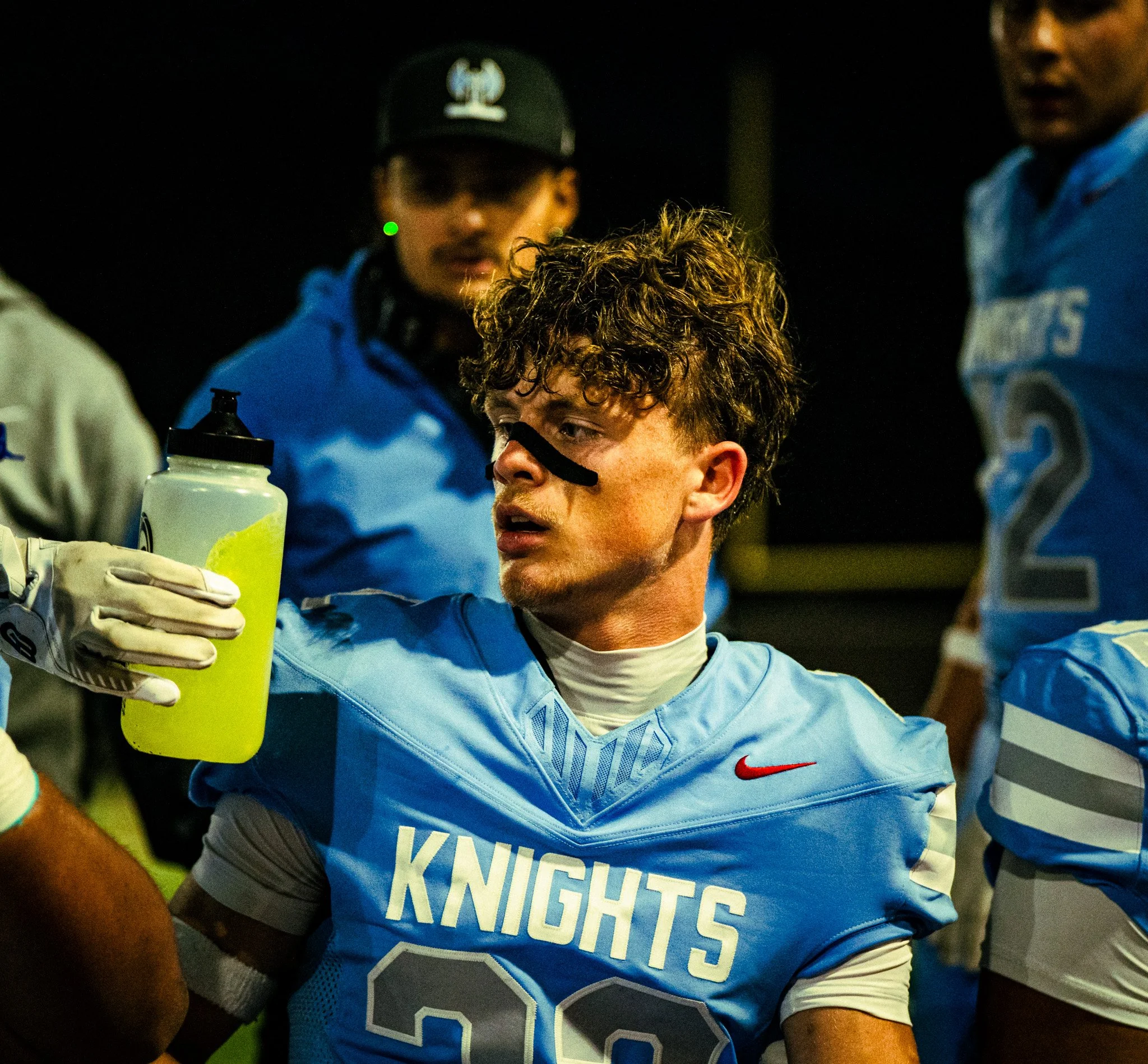 A football player with curly hair and black eye black is holding a water bottle, surrounded by teammates, during a night game.