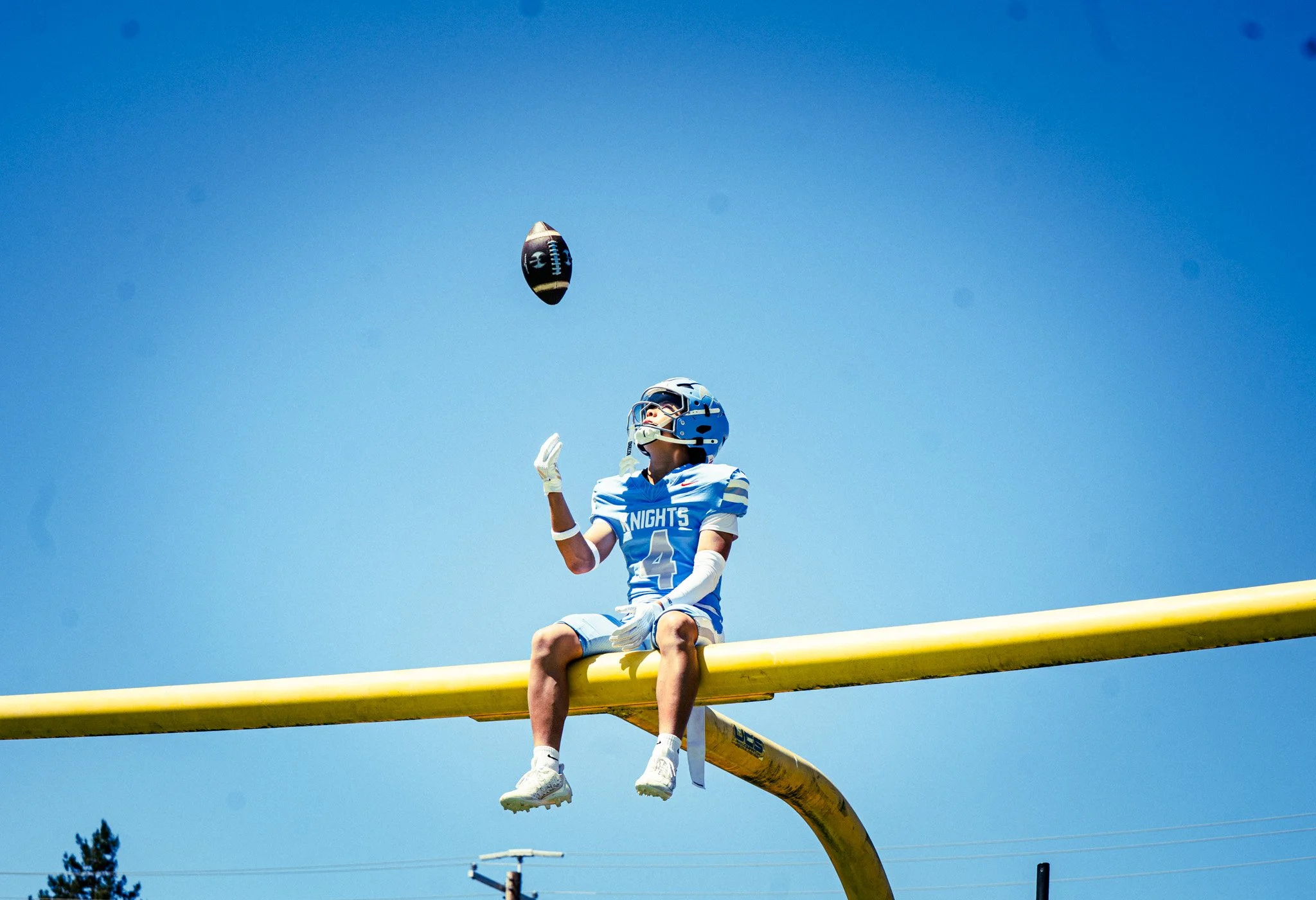 A football player in a blue and white uniform and helmet sitting on a yellow goal post, throwing a football in the air against a clear blue sky.
