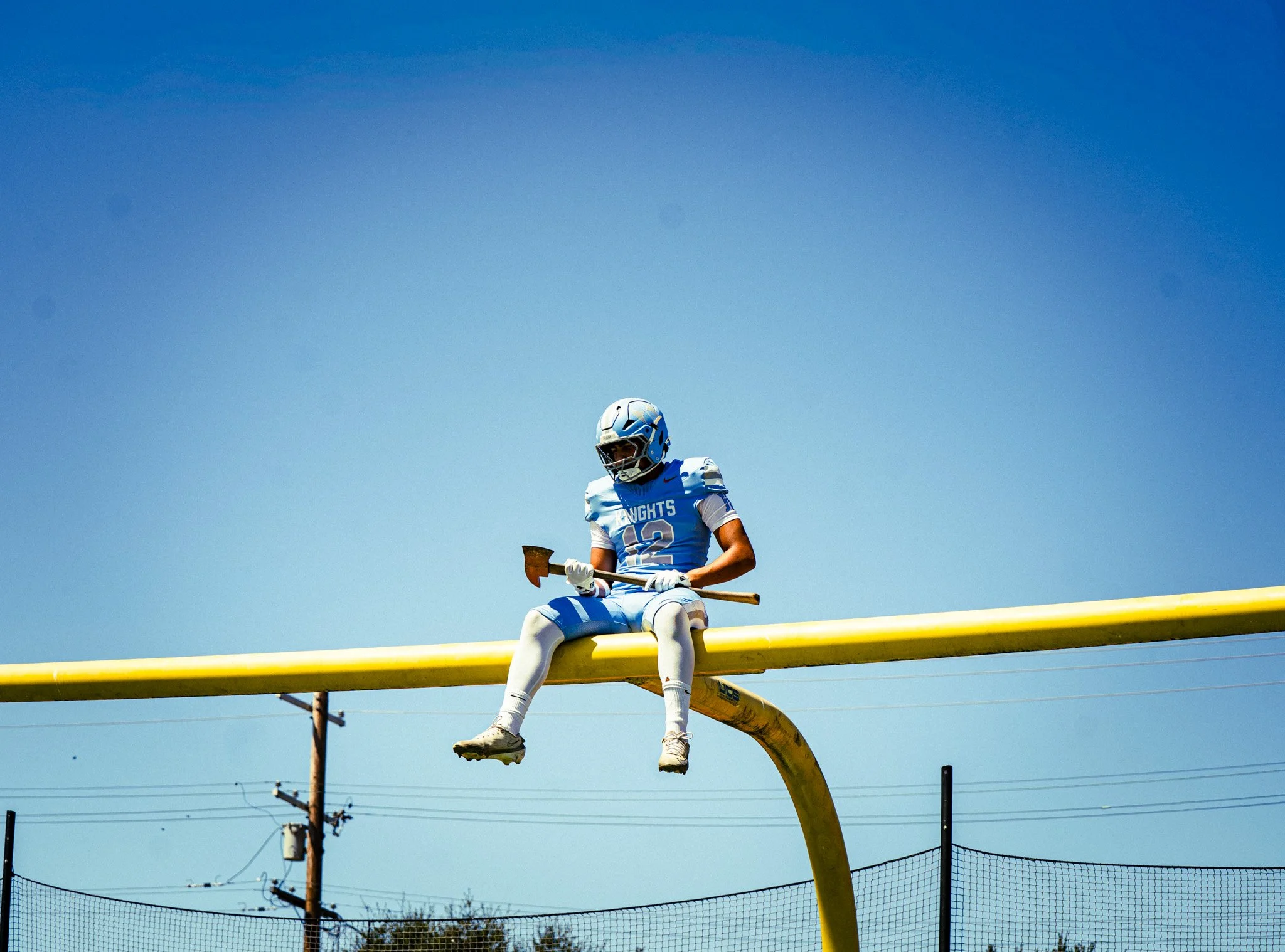 Football player wearing a blue and white uniform and helmet, sitting on a yellow goalpost crossbar under a clear blue sky.