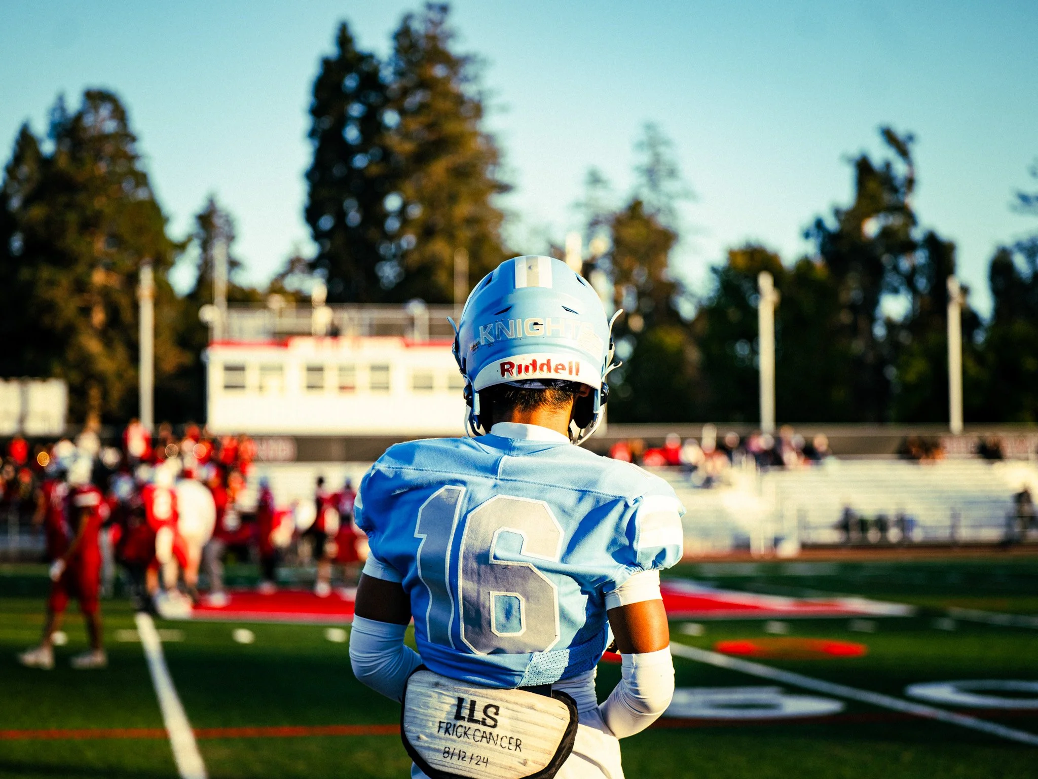 A football player wearing a blue and white uniform and helmet stands on the field during a game, with a crowd and trees in the background.