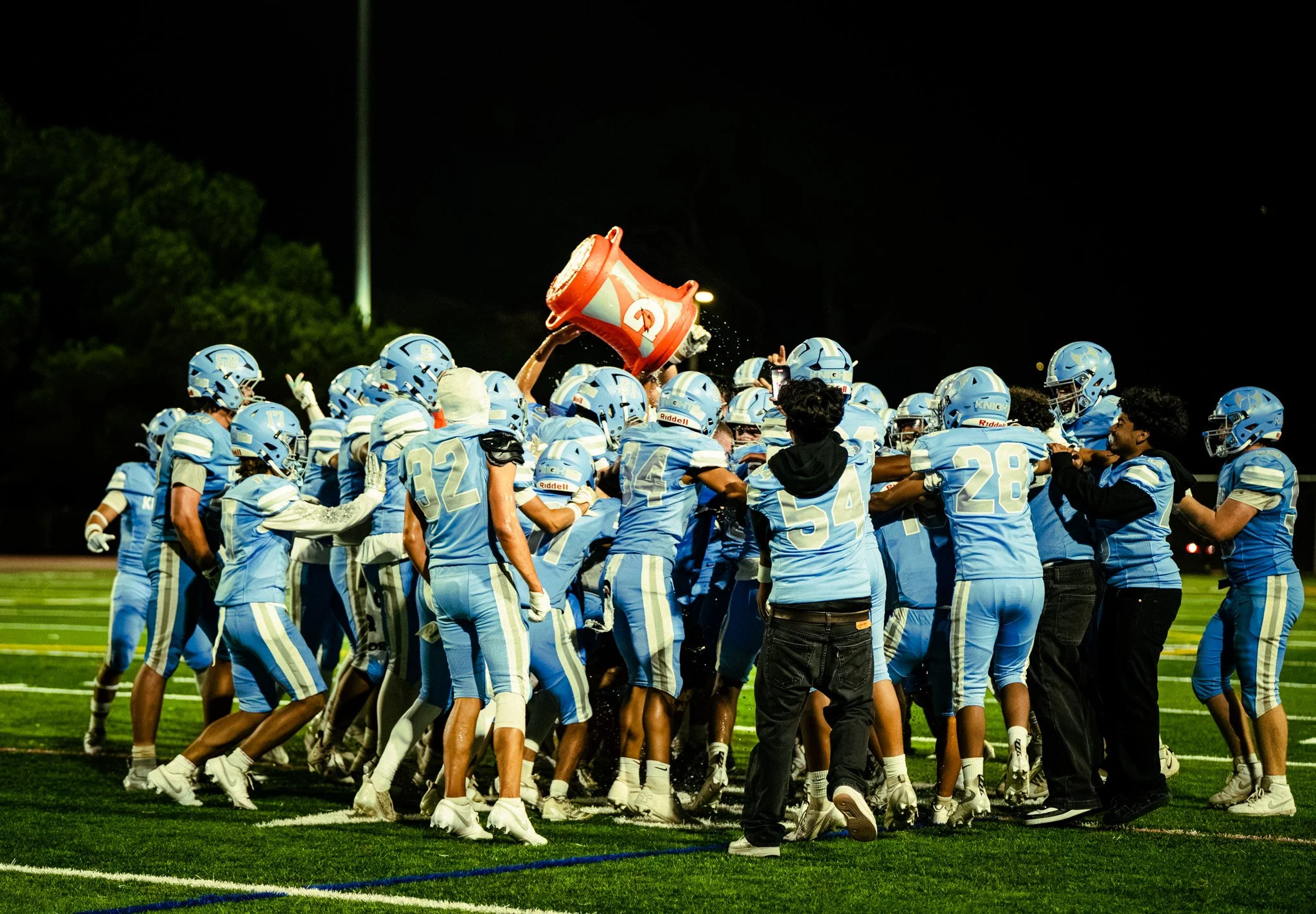 A group of football players in blue uniforms celebrating on the field at night, with one player holding a large Gatorade trophy above his head.