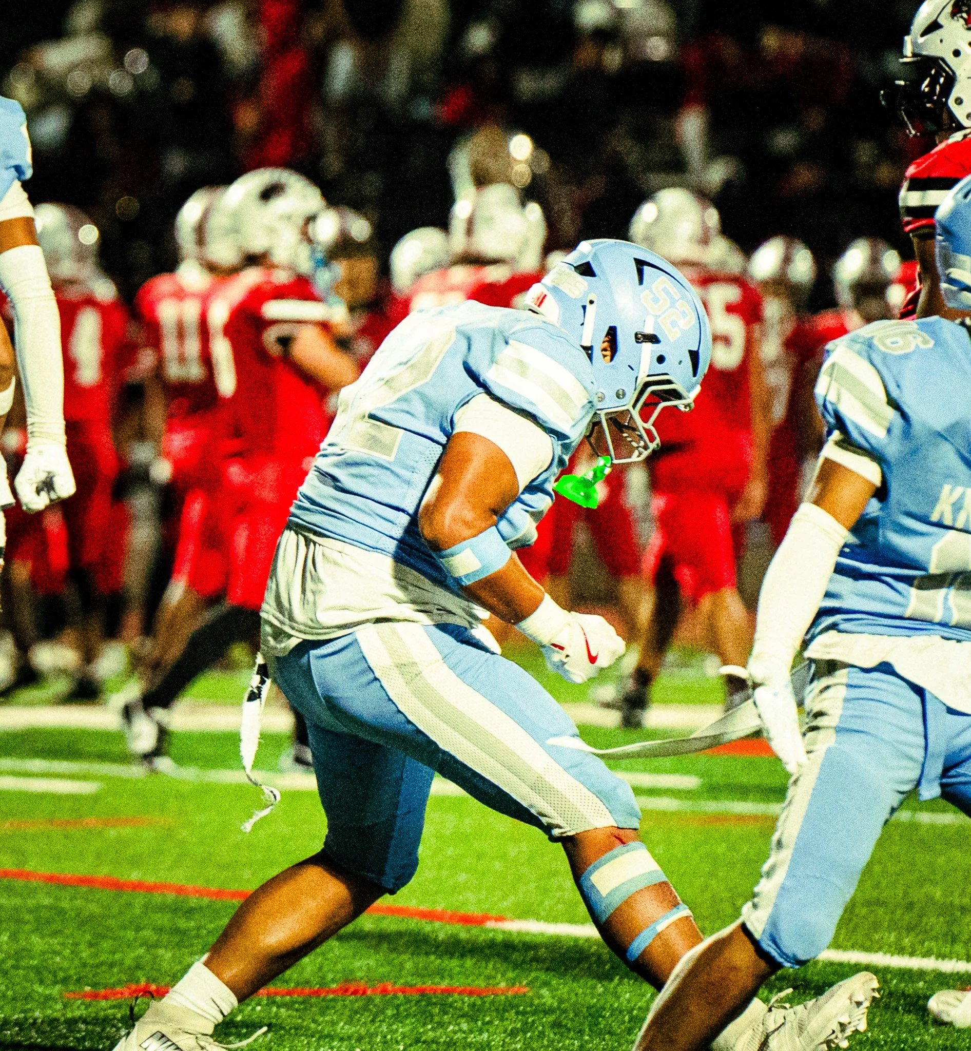 High school football game at night featuring players in blue and white uniforms facing an opposing team in red and white uniforms. Players are engaged in a play on the field, with focused attention on the game.