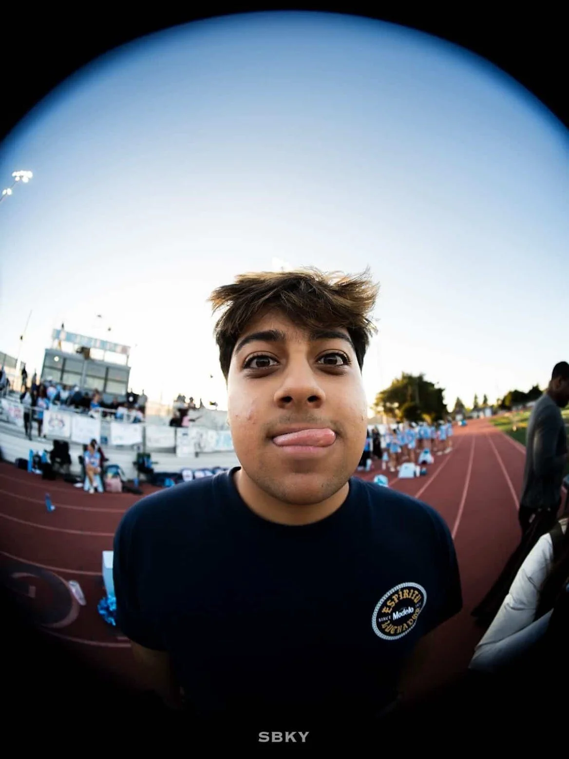 Close-up of a young man with brown hair making a face with his tongue out at a sports event on a track field, with people and event booths in the background.