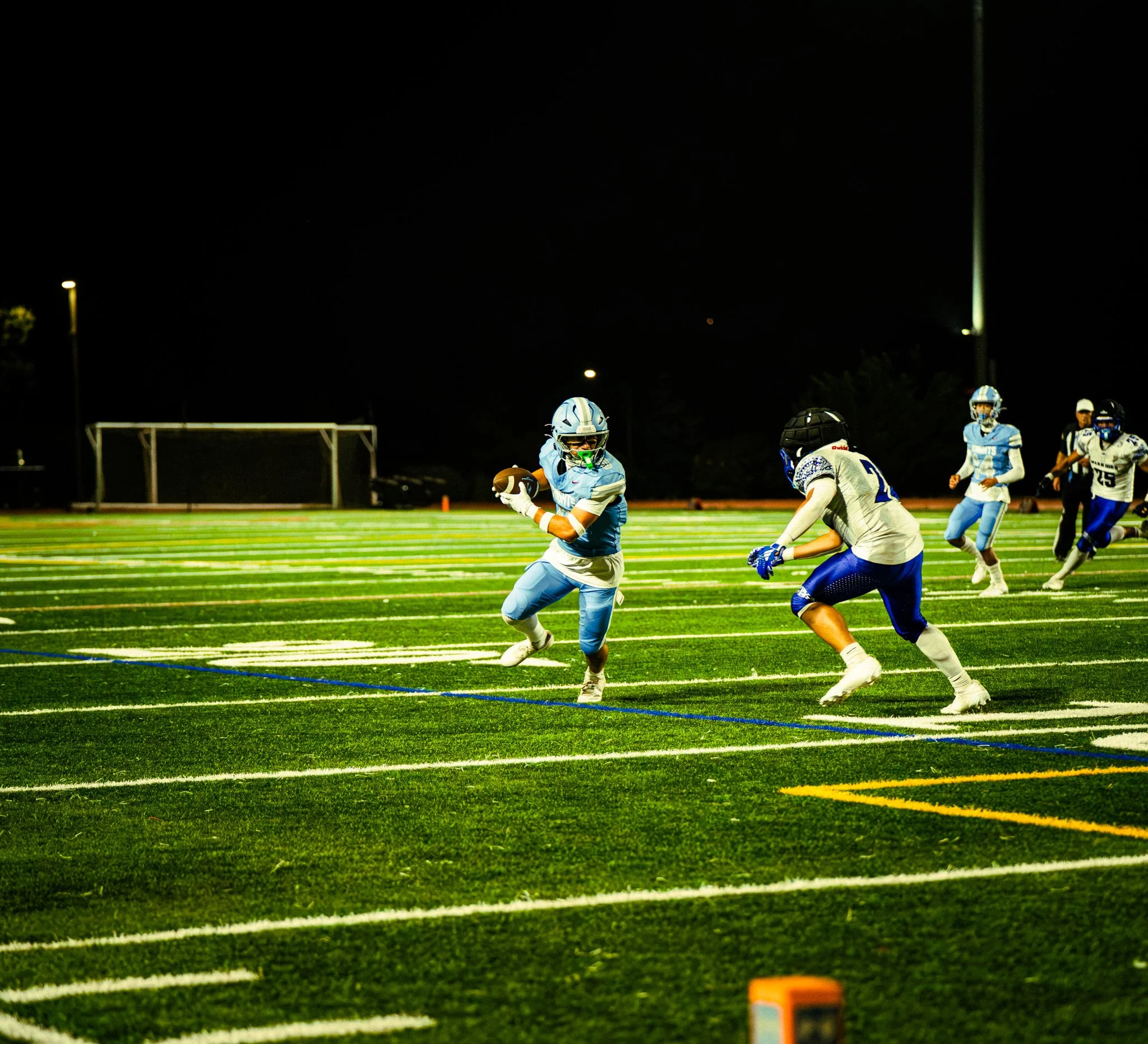 Nighttime football game on a bright green field with players in blue and white uniforms, some wearing helmets, and a dark sky overhead.