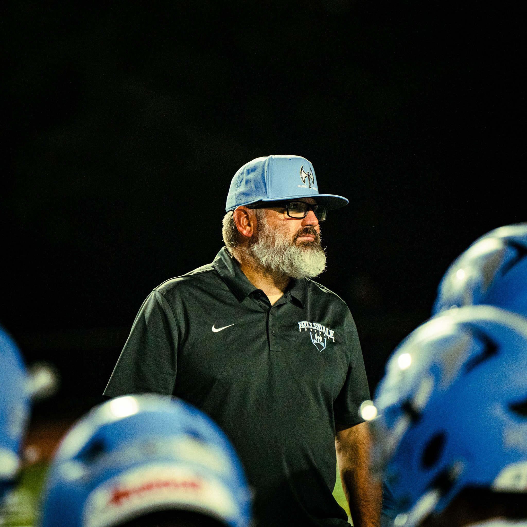 Image of a man with glasses and a gray beard wearing a light blue cap and black shirt with a logo, seen from a low angle at night, with a dark sky and some lit objects in the foreground.