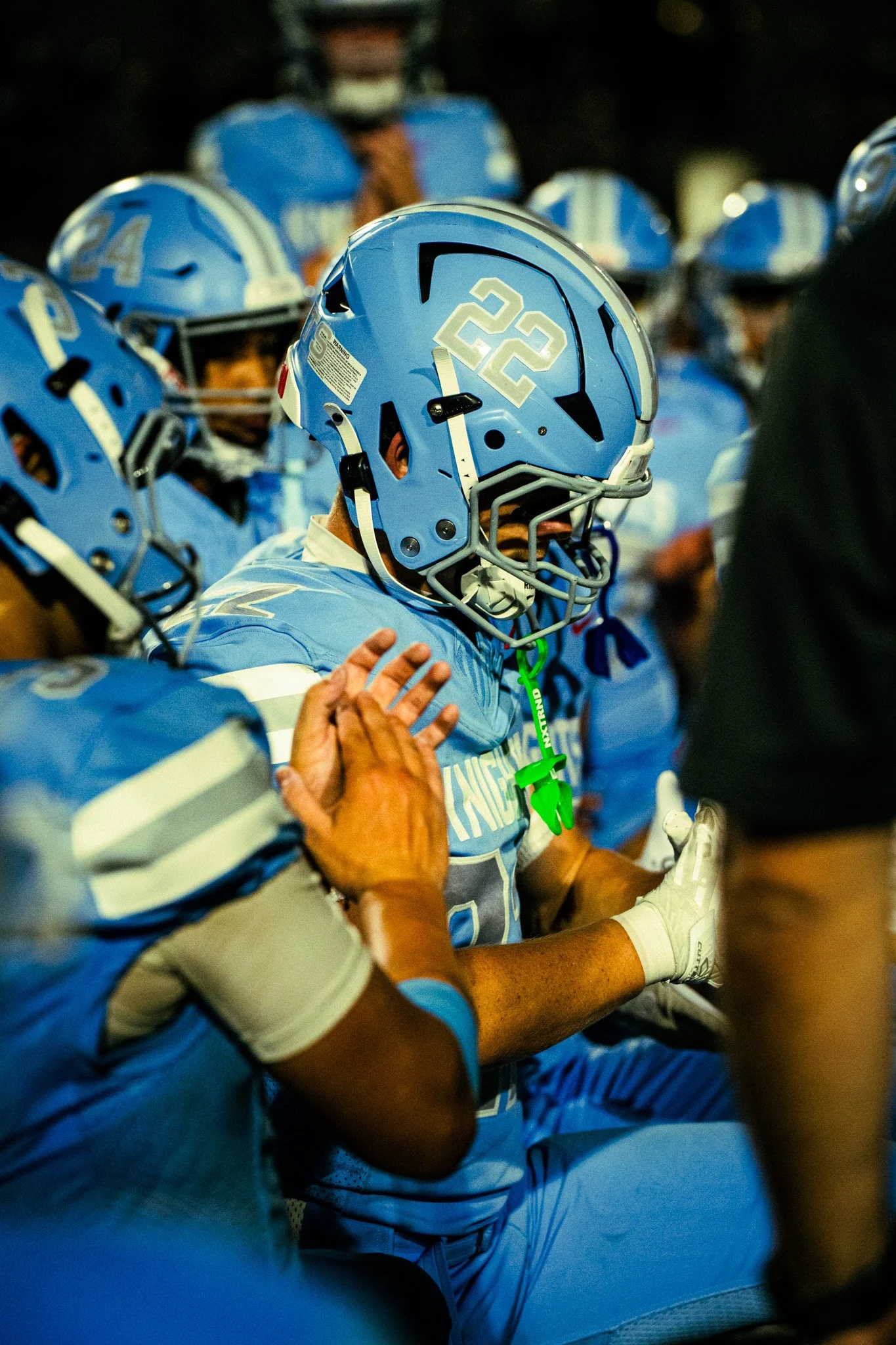 American football players in blue uniforms with helmets, gathered in a huddle, with one player in focus and others in background.