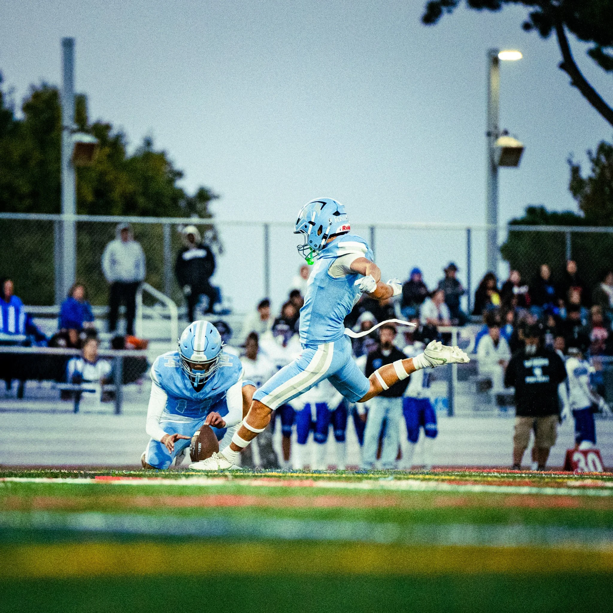 A football player in a light blue uniform and helmet is on a football field during a game, with spectators in the background.