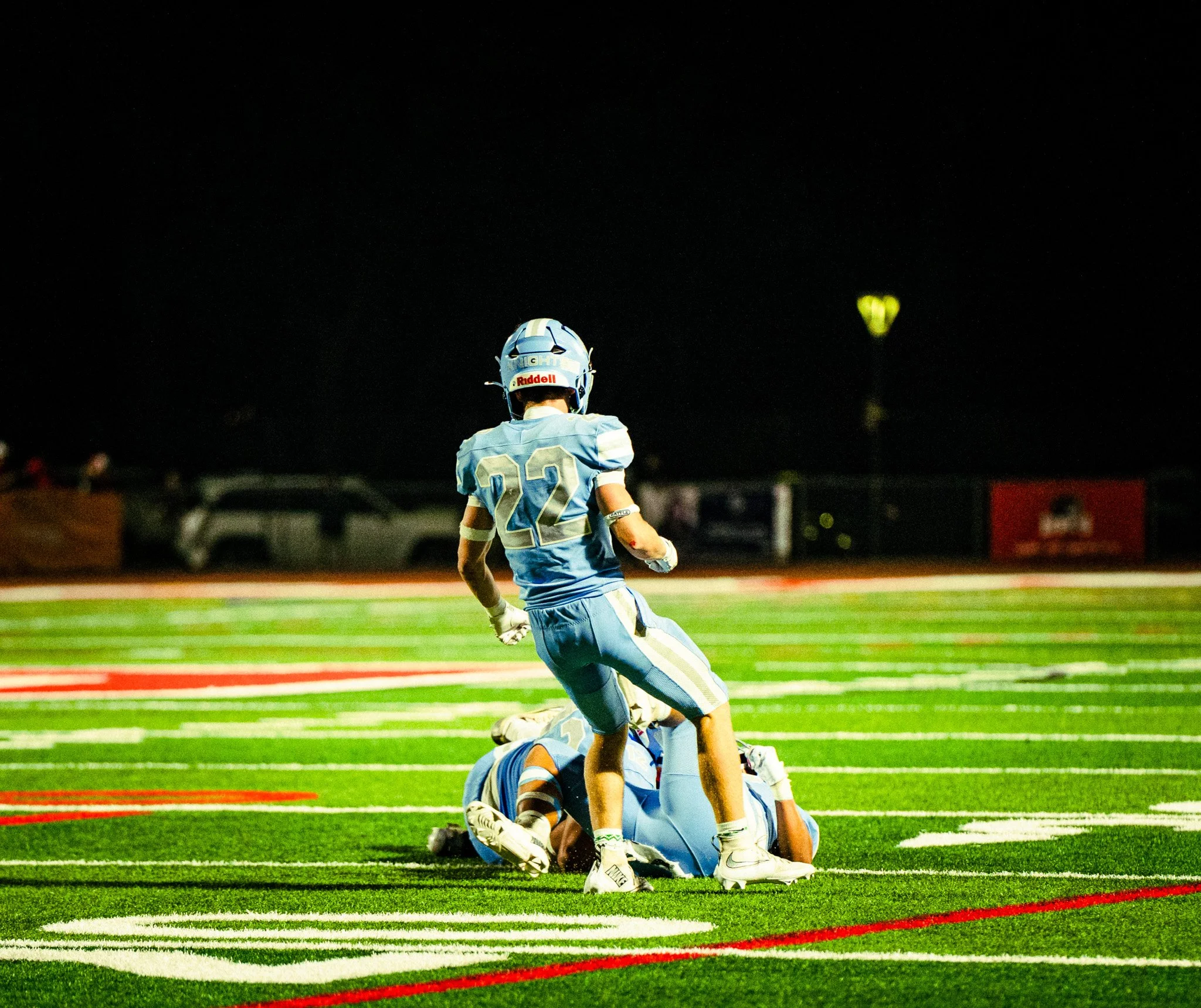 A football game at night with a player in a light blue uniform and helmet, number 22, standing on the field. The player appears to be celebrating on top of a teammate who is lying on the ground. The field has green turf with red and white yard lines,