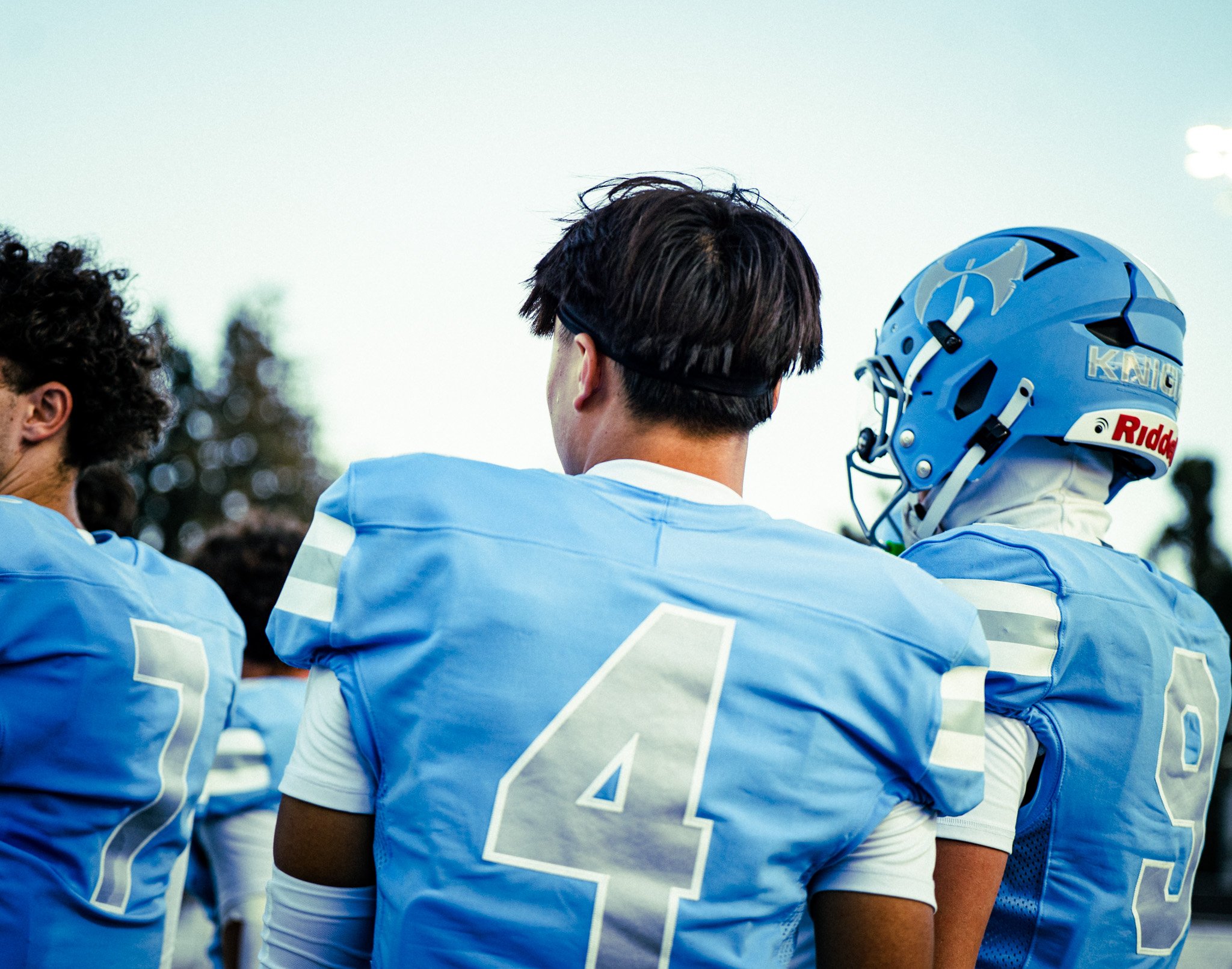 A group of football players in blue and white uniforms huddle together, with one player wearing a helmet, during a game or practice outdoors.