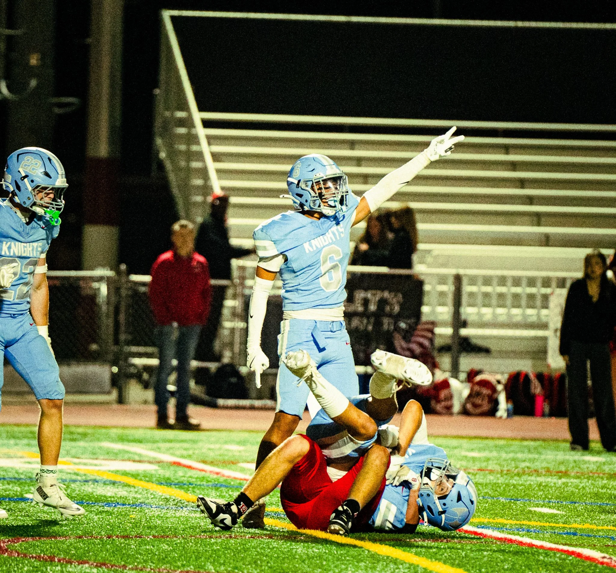 A football game scene at night showing players on the field. One player in a red jersey is on the ground, while other players in light blue jerseys are nearby; one with a glove and helmet, reaching out. The photo captures a moment of action, with spe