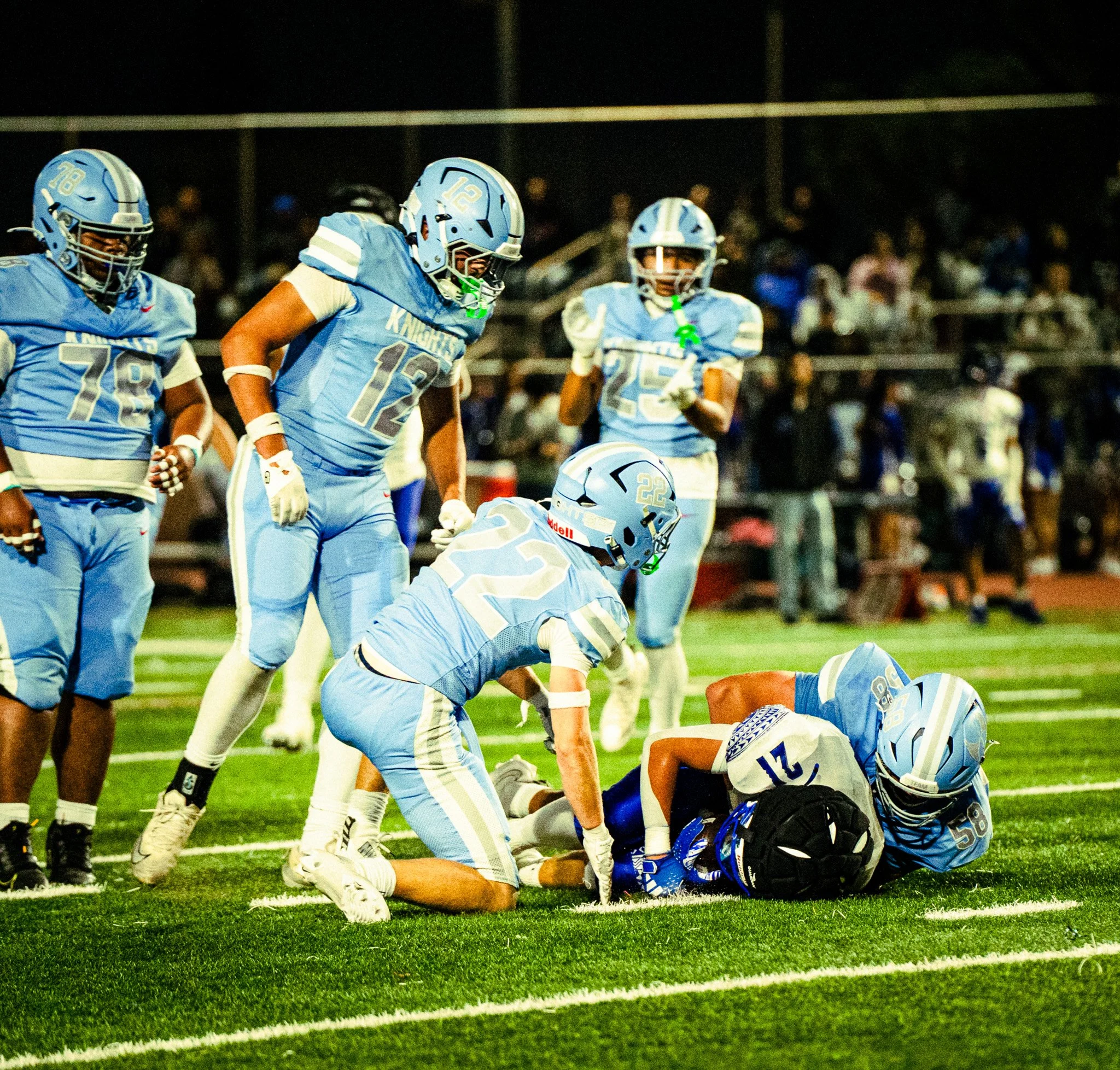 Football players in blue uniforms celebrate a play on the field at night, with a crowd in the background.