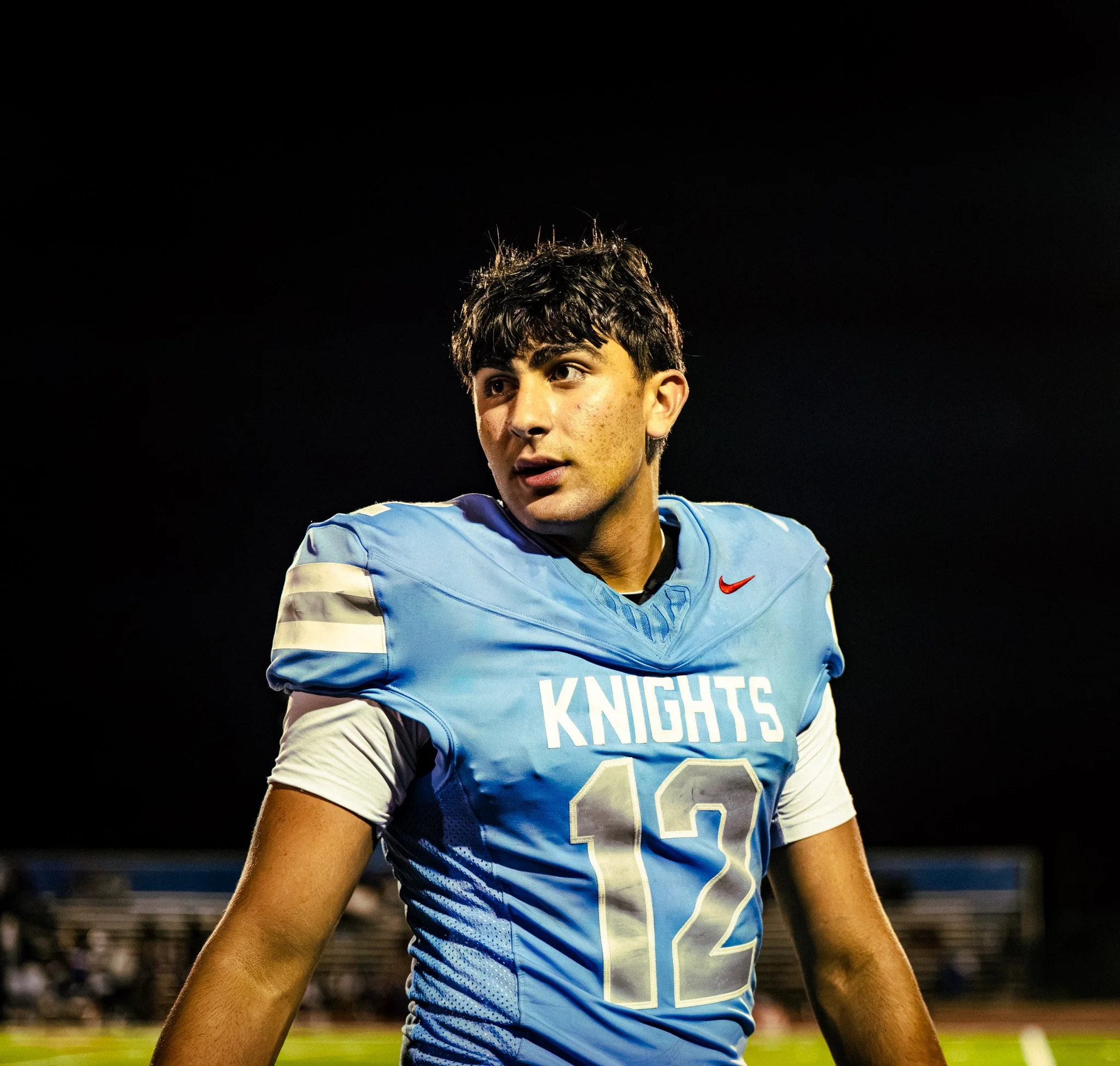 Close-up of a young male football player with short dark hair, wearing a light blue jersey with the word 'KNIGHTS' and the number 22, standing on a football field at night.