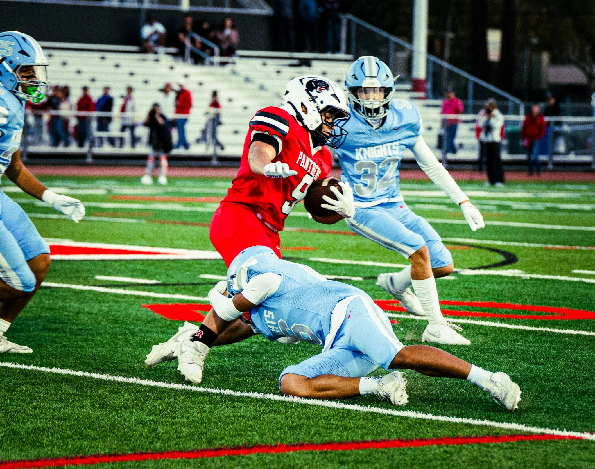 An American football game with two teams, one in red uniforms and one in light blue uniforms. A player in red holds the football and is tackled by a player in light blue who is on the ground. Other players and spectators are visible in the background