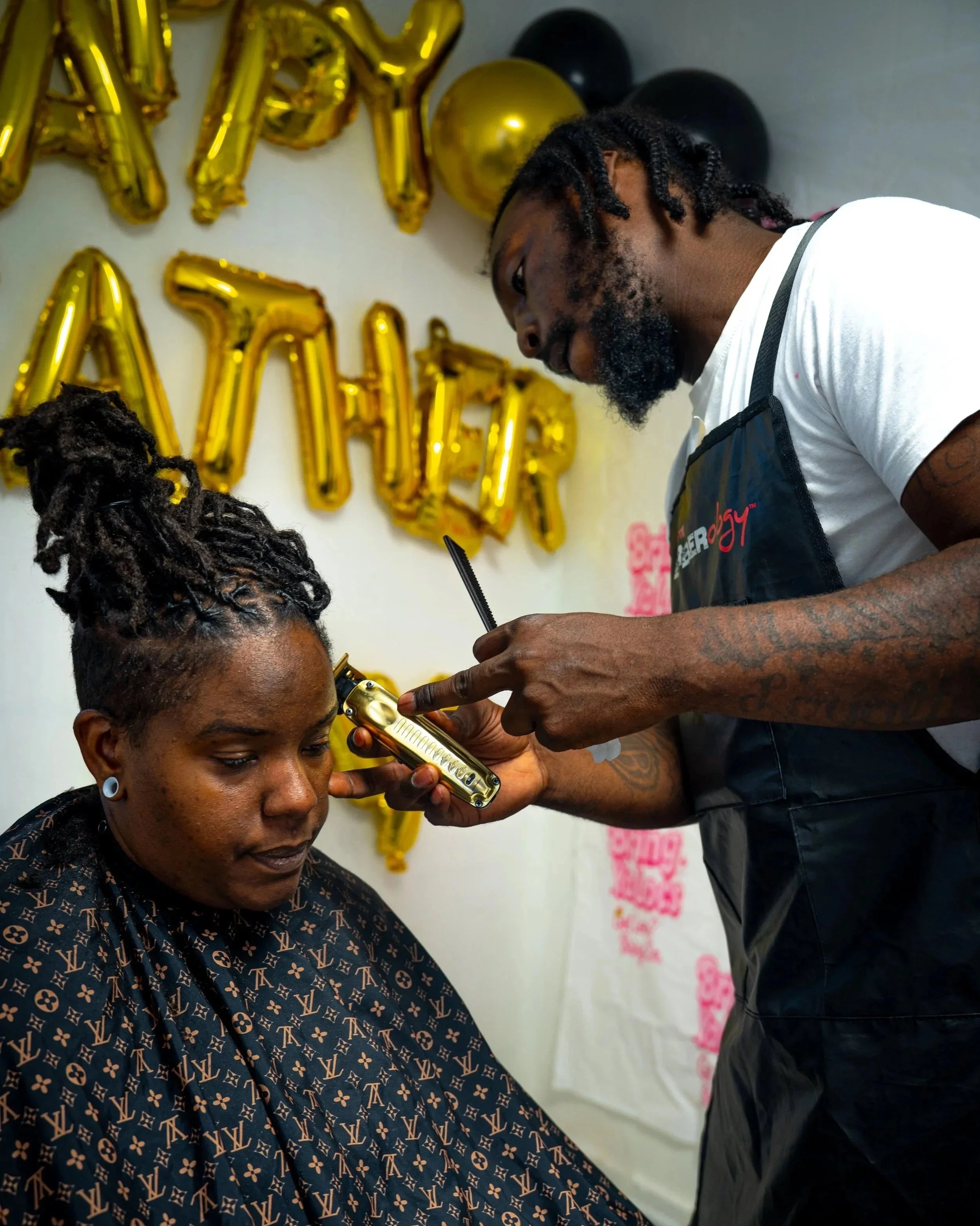 A man with dreadlocks gives haircuts to women in a celebration setting with gold and black balloons and 'HAPPY BIRTHDAY' balloons in the background.