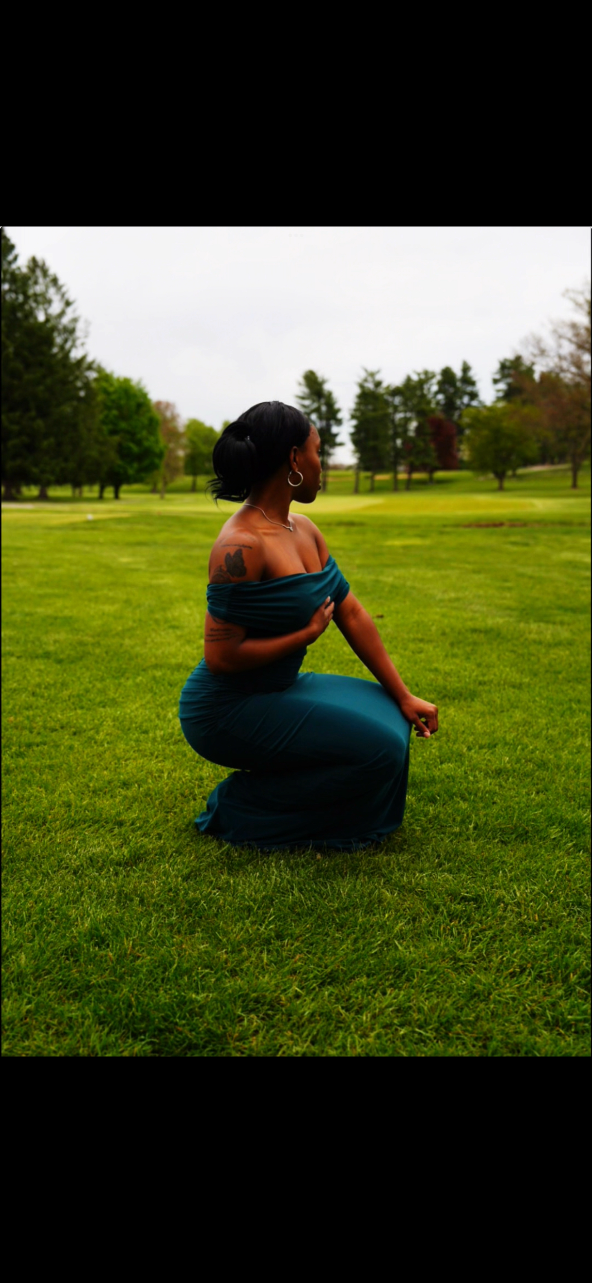 A woman wearing a teal off-shoulder dress, with tattoos on her arm, kneeling on a grassy field in a park with trees in the background.