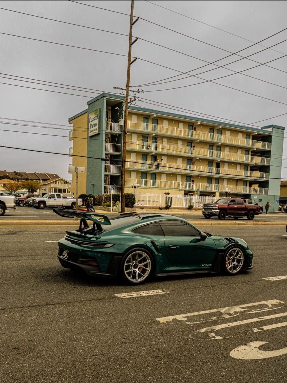 A green sports car with a large rear wing is parked on the street in front of a yellow and blue multi-story apartment building named Coastal Palms. Several cars and pedestrians are visible on the road and sidewalk.