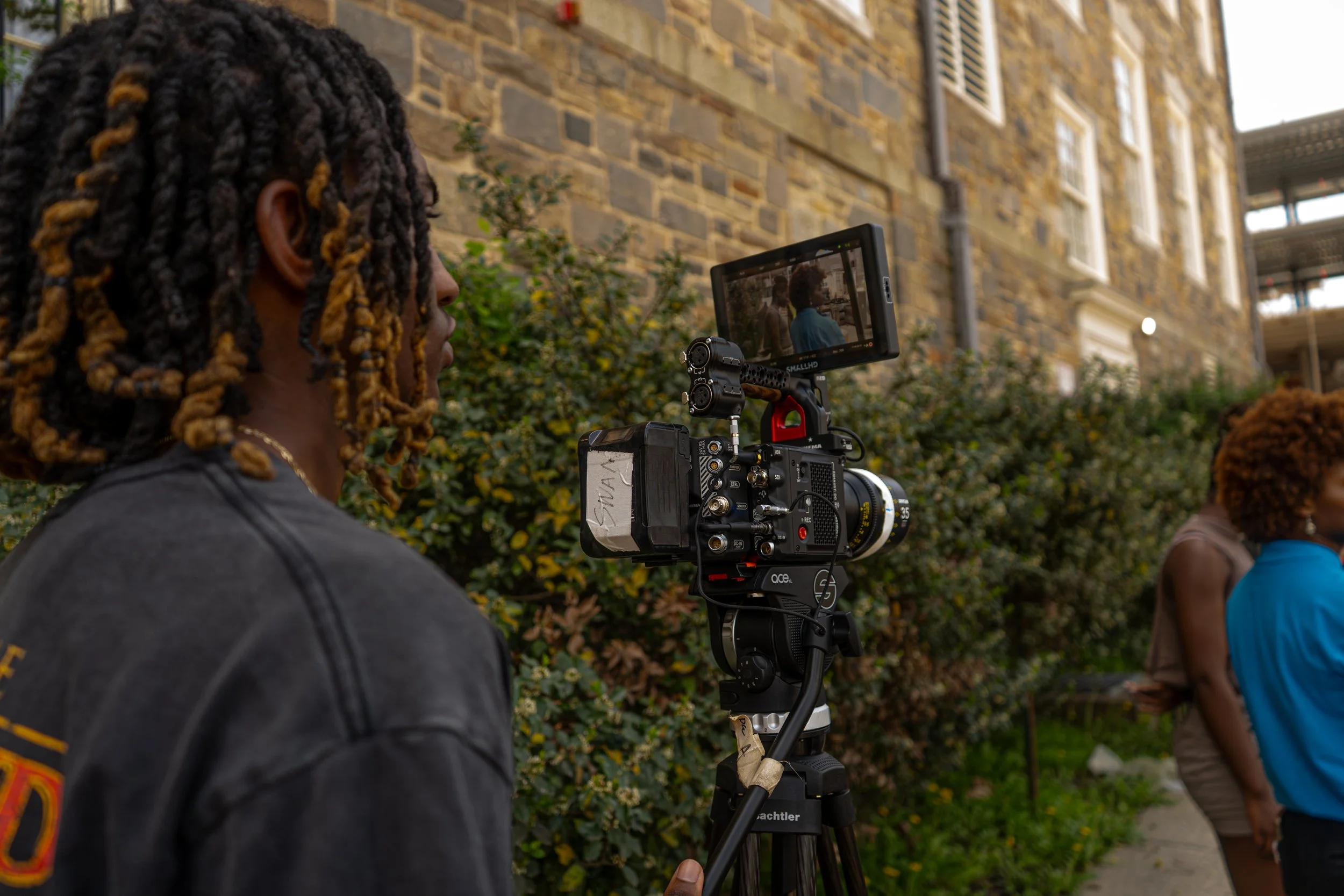 A person with dreadlocks is filming with a professional camera mounted on a tripod outdoors, with a brick building and greenery in the background.