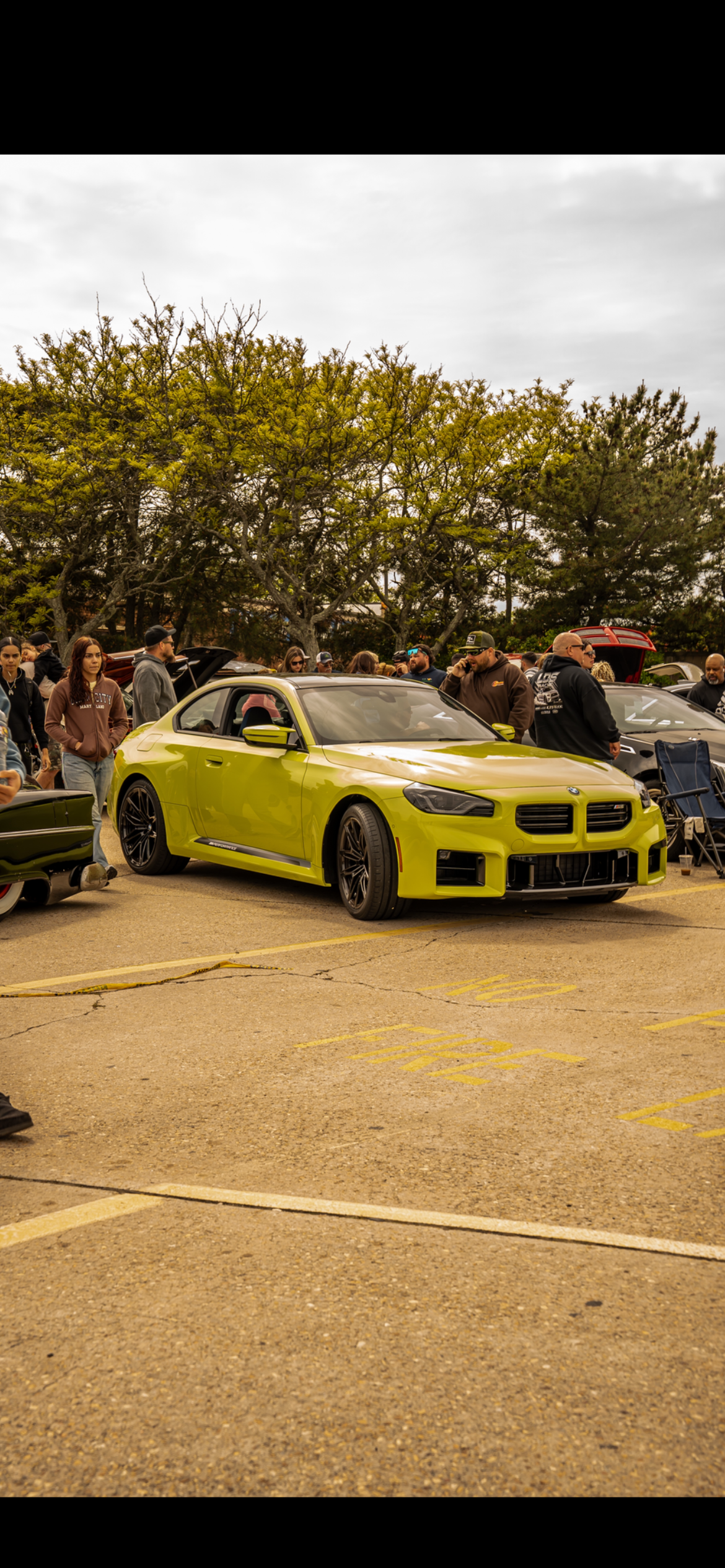 A bright green sports car parked at a car show with a crowd of people around it, some looking at other vehicles, in an outdoor lot under cloudy skies.