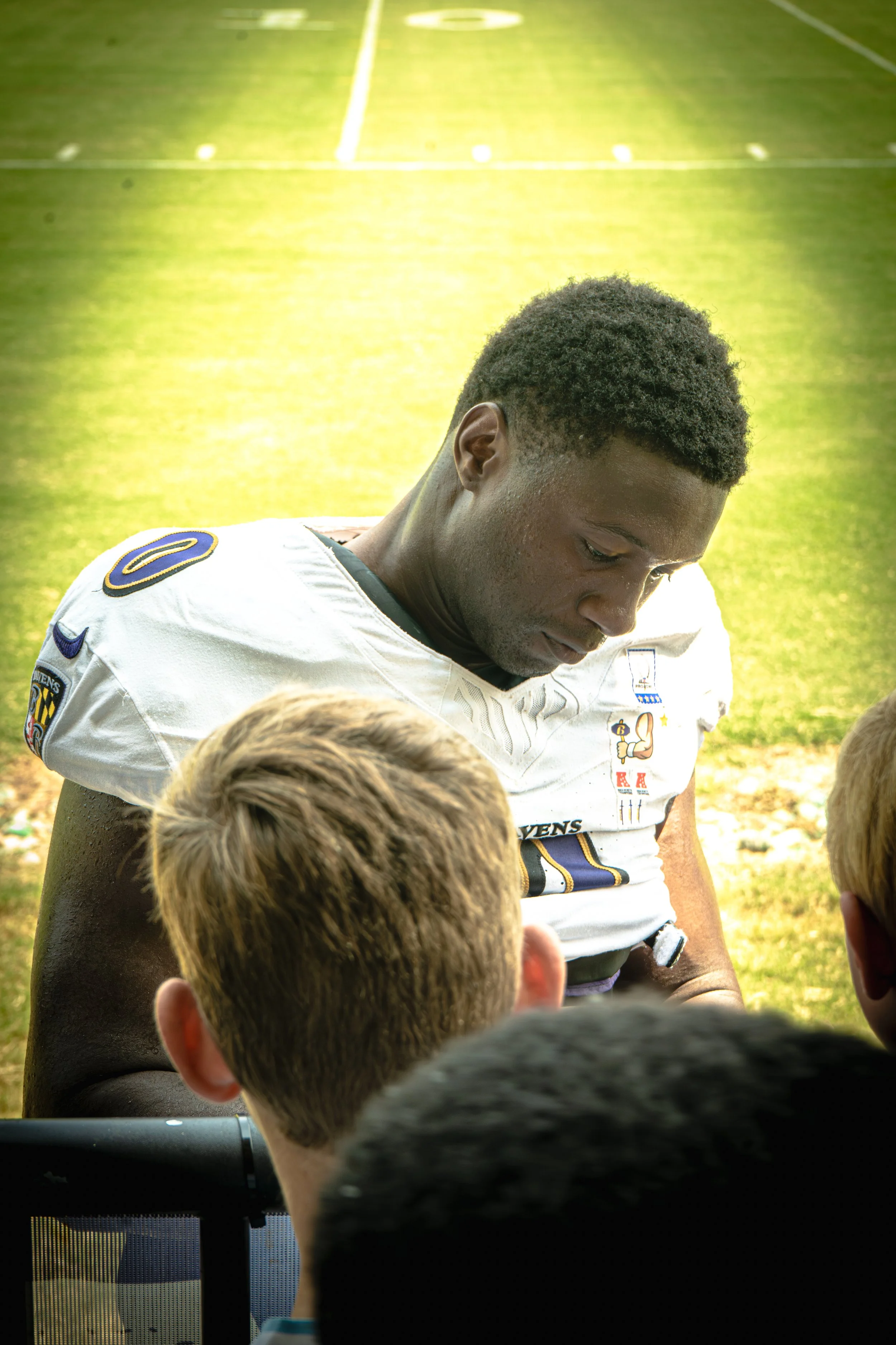 A football player is sitting on the field wearing a white jersey with various patches and a number 8 on the shoulder. Several children with blond hair are gathered around him, some looking up at him. The scene is outdoors on a grassy field, possibly after a game or practice.