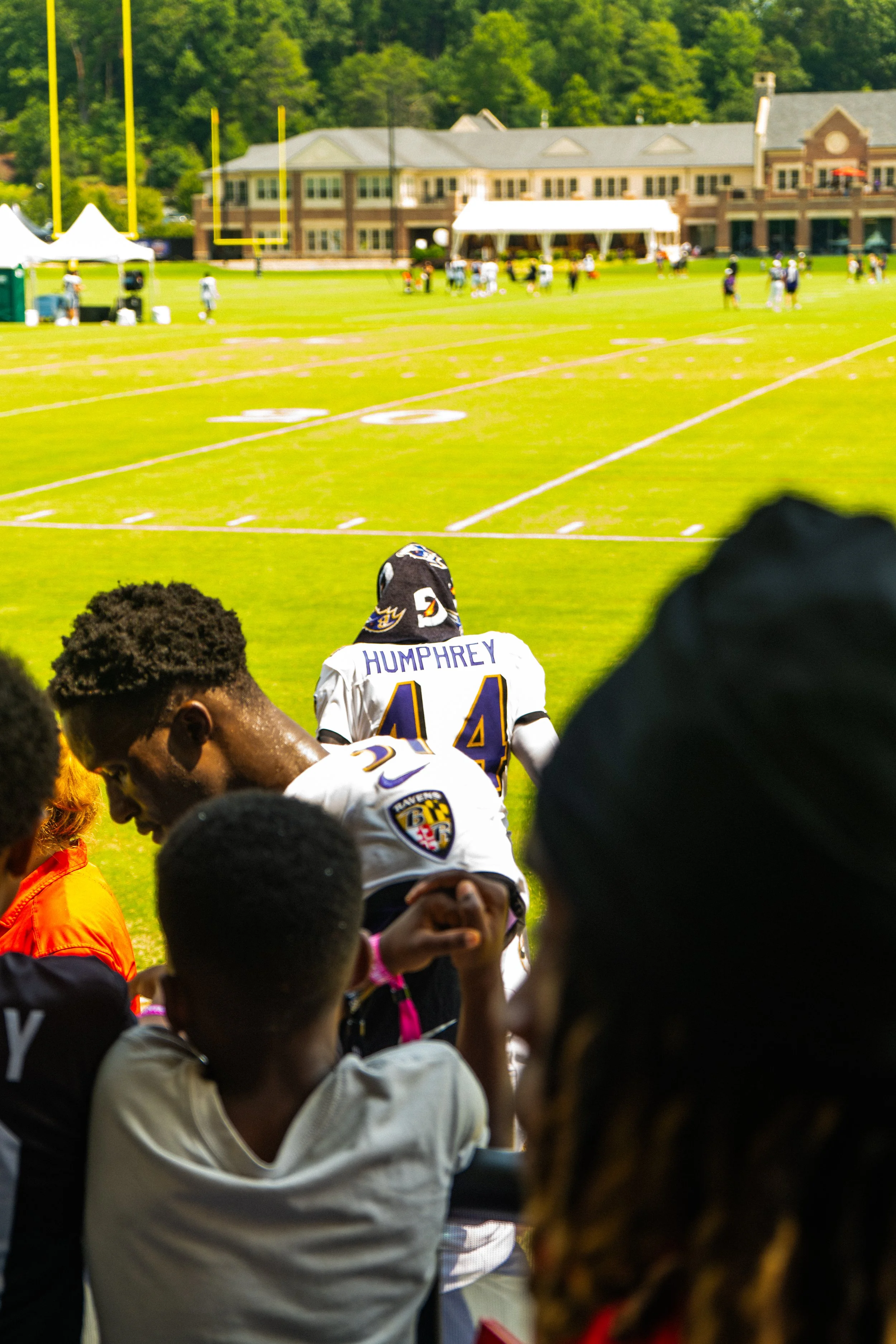 Children and adults at an outdoor football game, with players in football gear on the sideline and a building in the background.