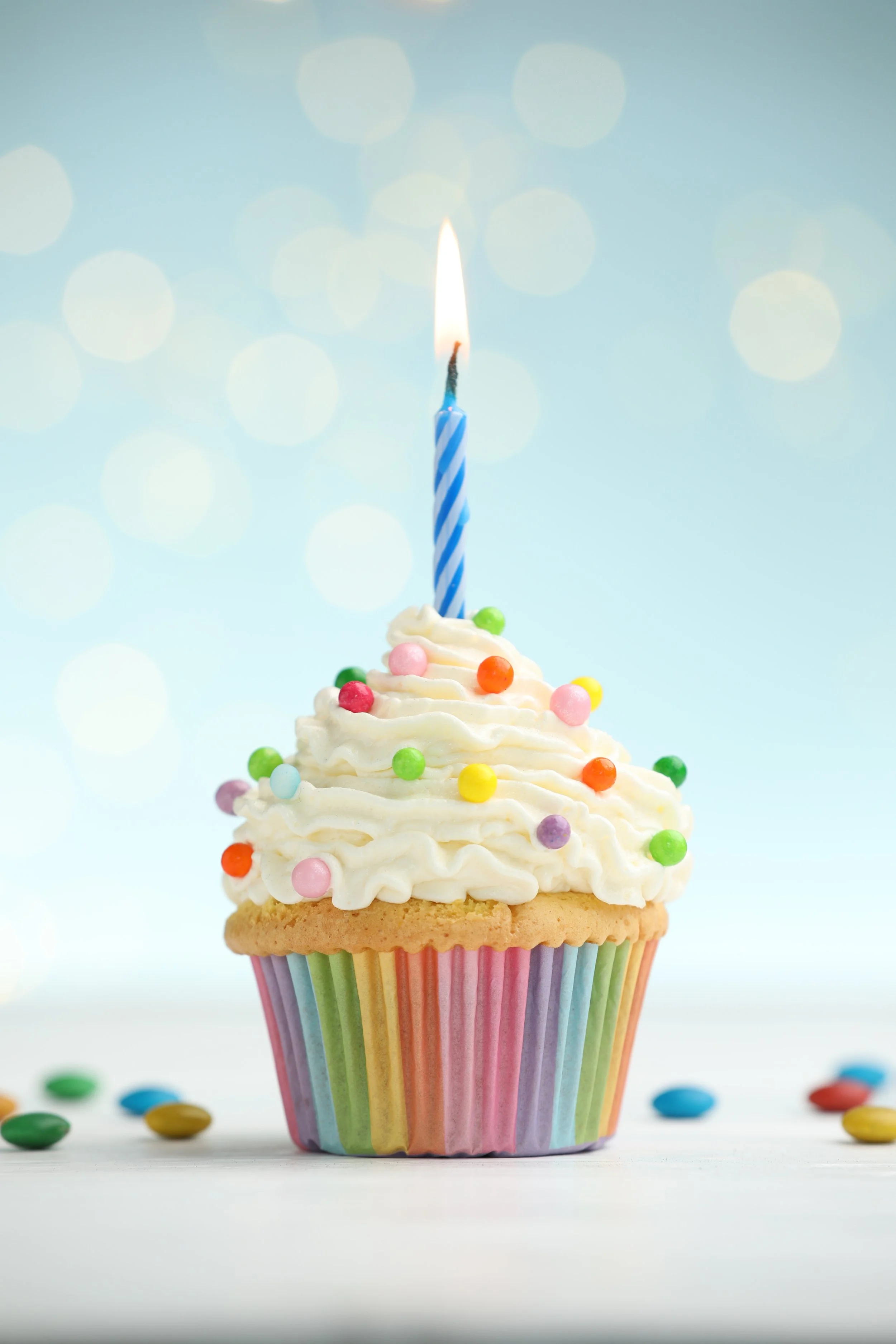 Colorful birthday cupcake with white frosting, rainbow sprinkles, and a lit blue striped candle, set against a light blue background with bokeh light effects.