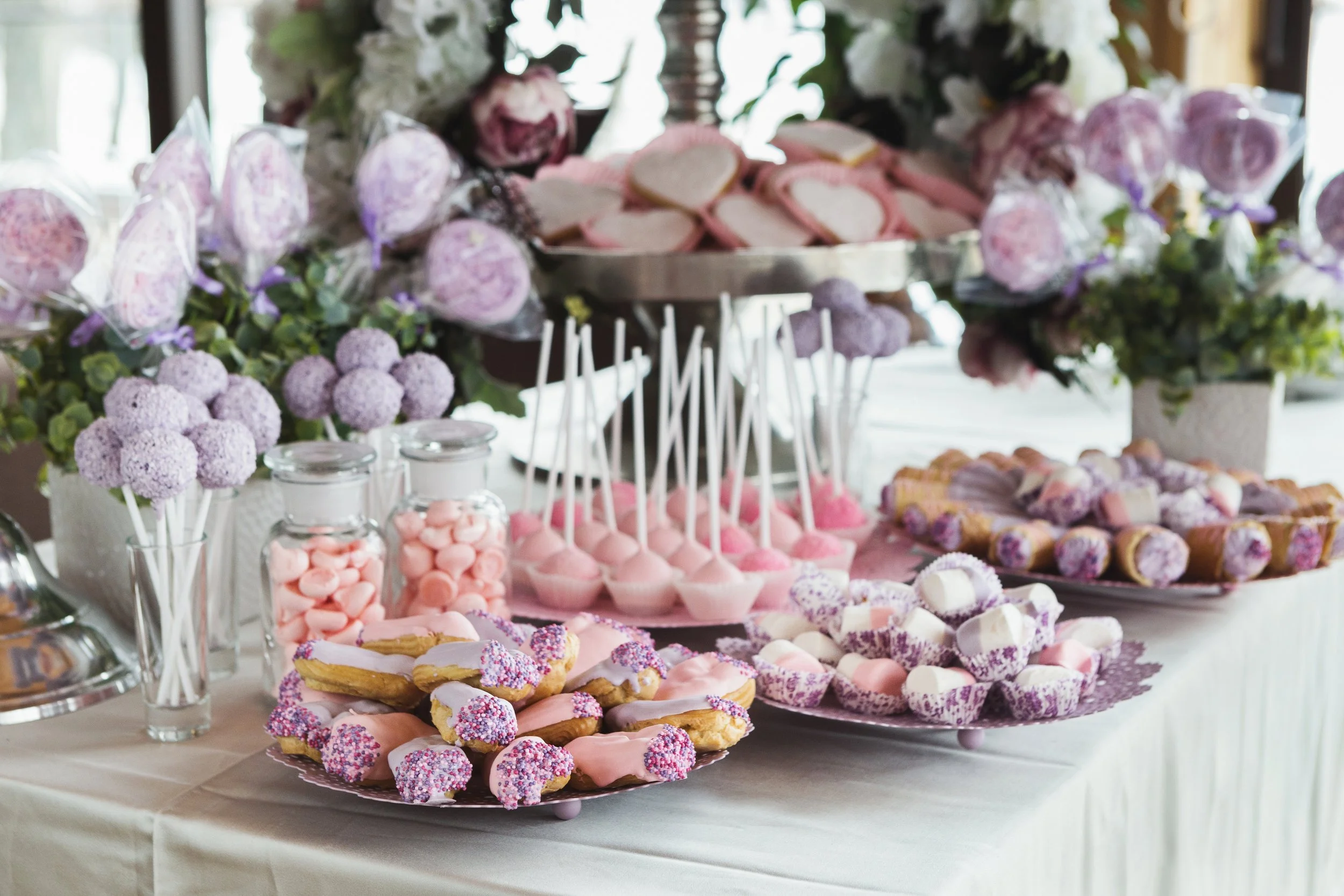 A table decorated with pink and purple desserts, including cupcakes, cookies, cake pops, and marshmallows, surrounded by floral arrangements with purple flowers and greenery.