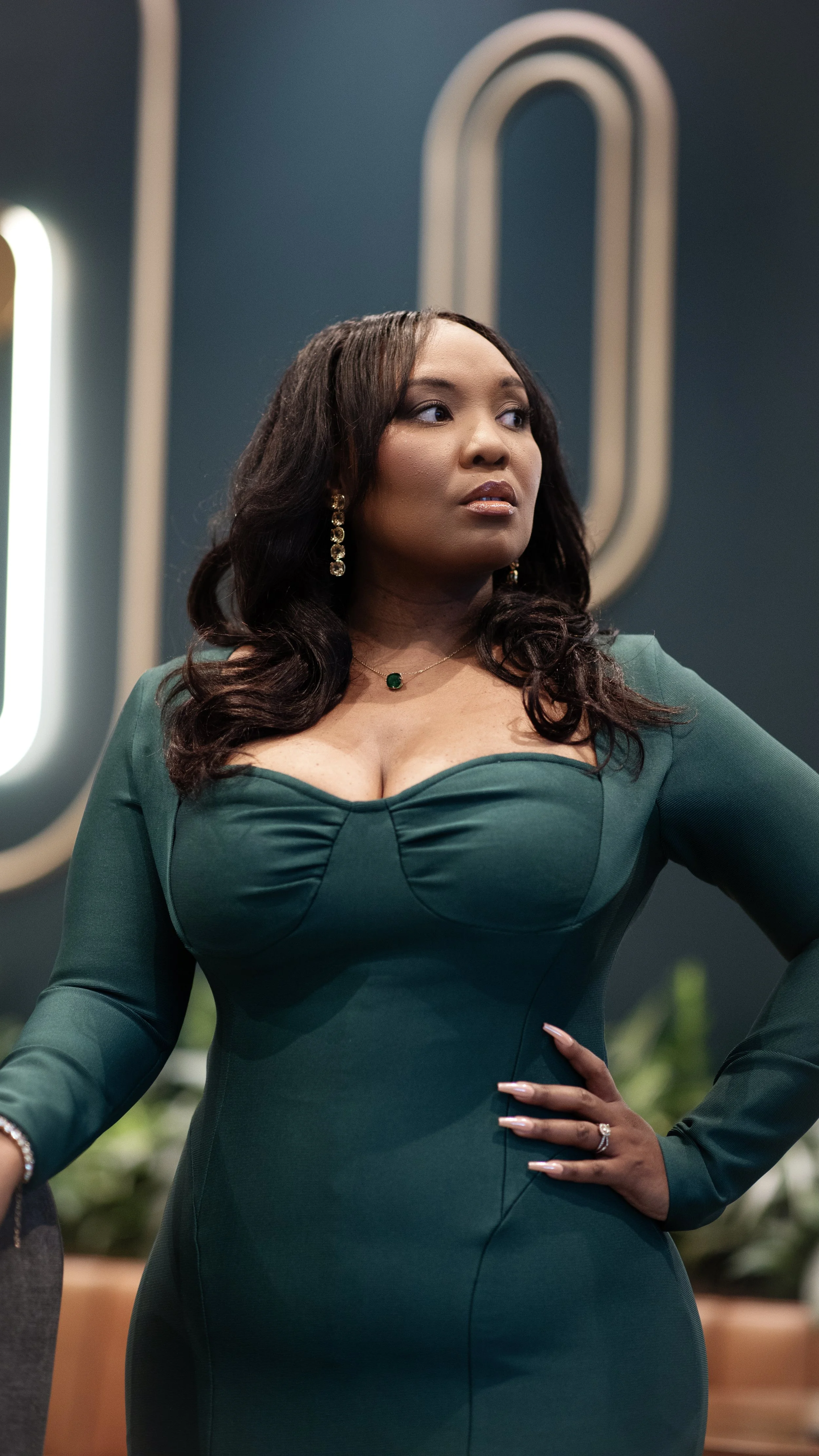 A woman wearing a green dress with a plunging neckline and long sleeves. She has long, dark, wavy hair, gold dangling earrings, a green pendant necklace, and a ring on her left hand. She is standing indoors with her right hand on her hip, and there is a neon sign in the background.