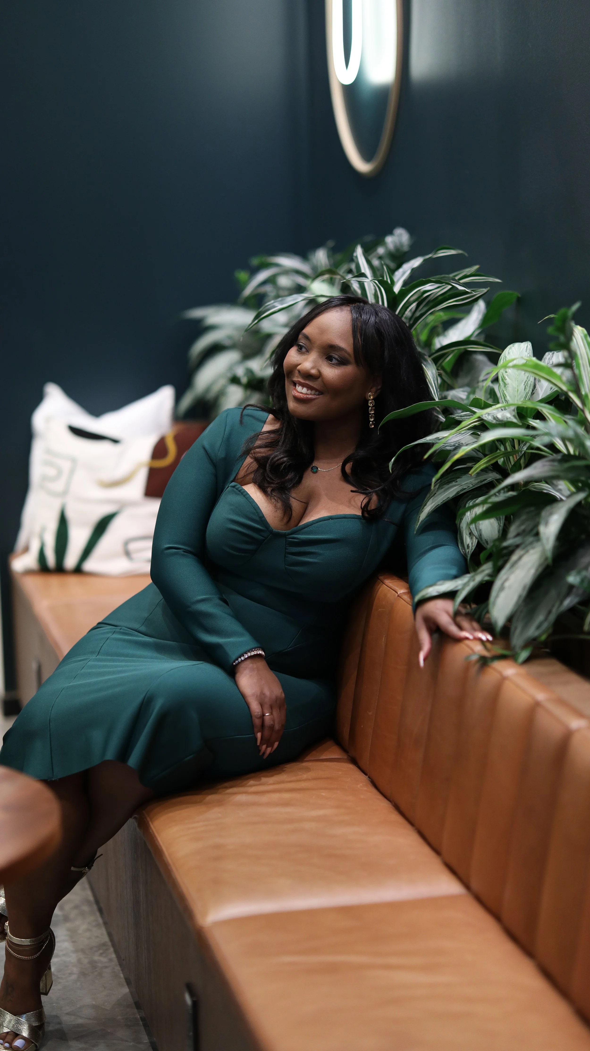 A woman in a green dress sitting on a leather bench surrounded by green plants.
