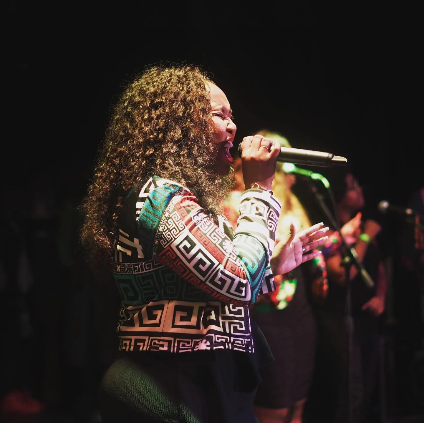 A woman singing into a microphone on stage with her eyes closed and hair in curls, wearing a colorful geometric patterned jacket, with other performers in the background.