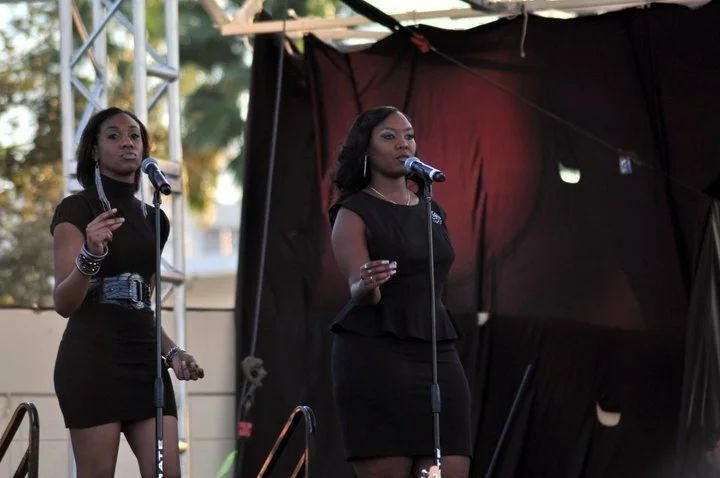 Two women speaking or singing on an outdoor stage with microphones, with a black backdrop and stage lighting.