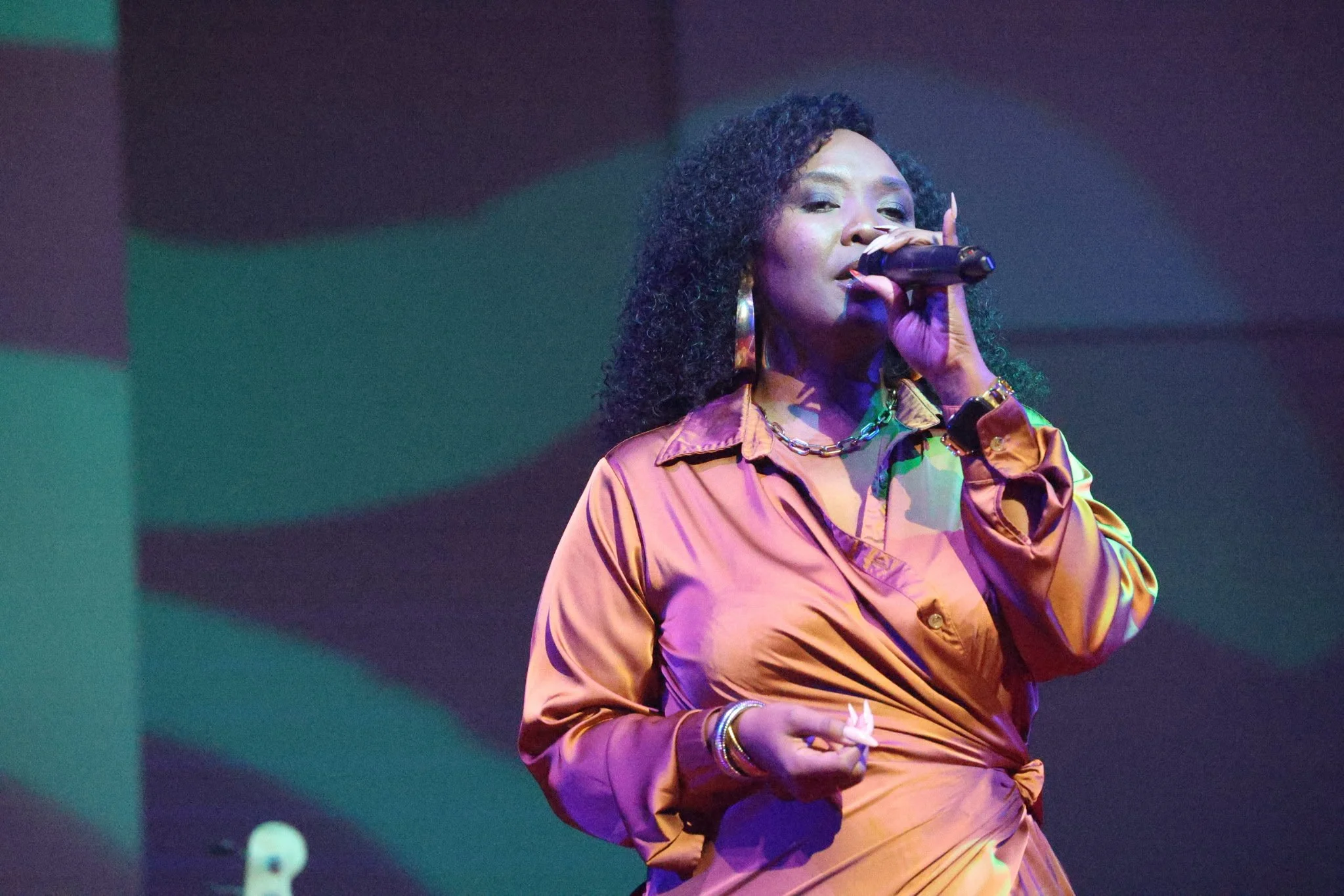Woman with curly hair singing into a microphone, wearing an orange satin shirt with jewelry, against a dark background.