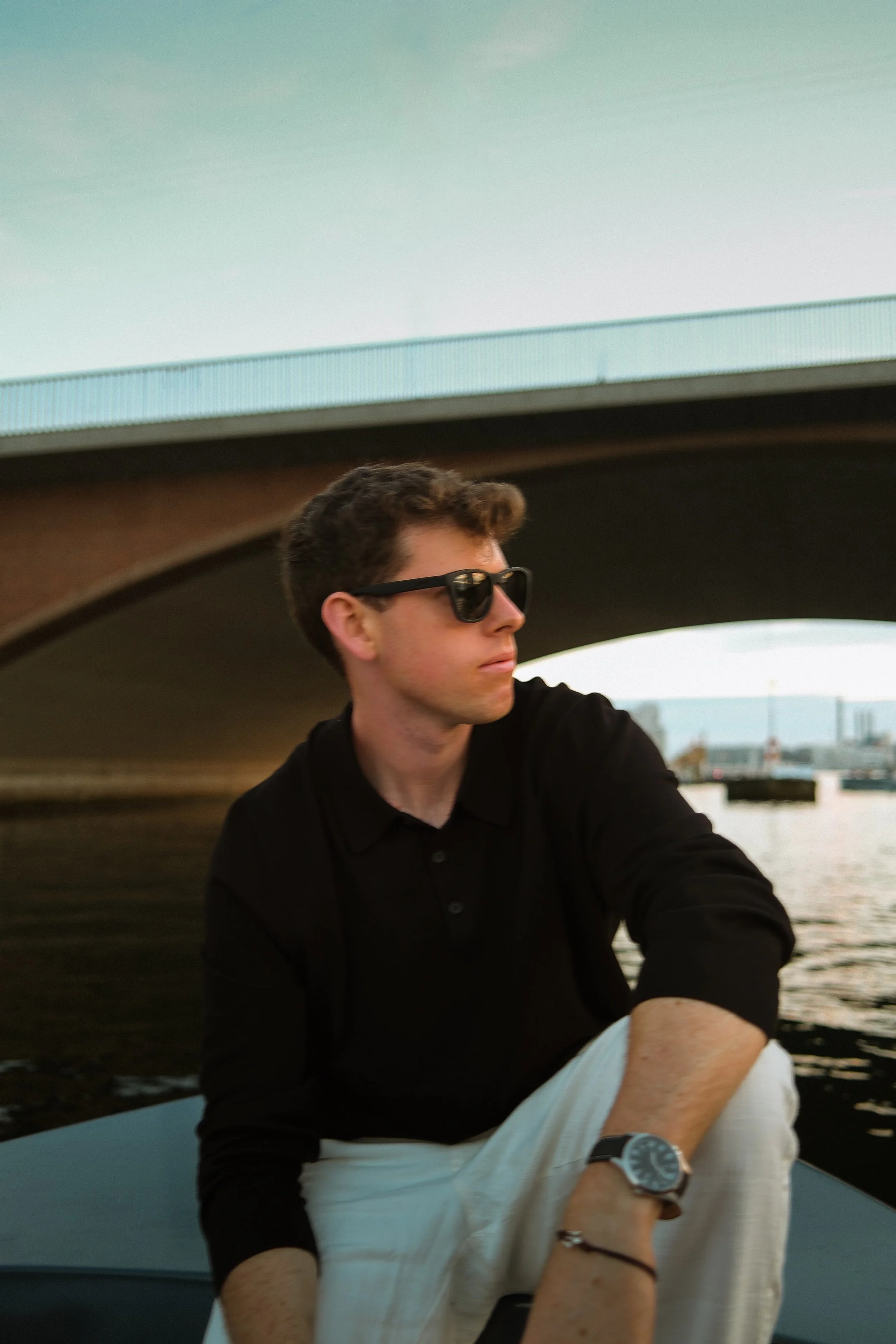 A young man with curly brown hair wearing black sunglasses and a black shirt, sitting near water under a bridge, with city buildings in the background.