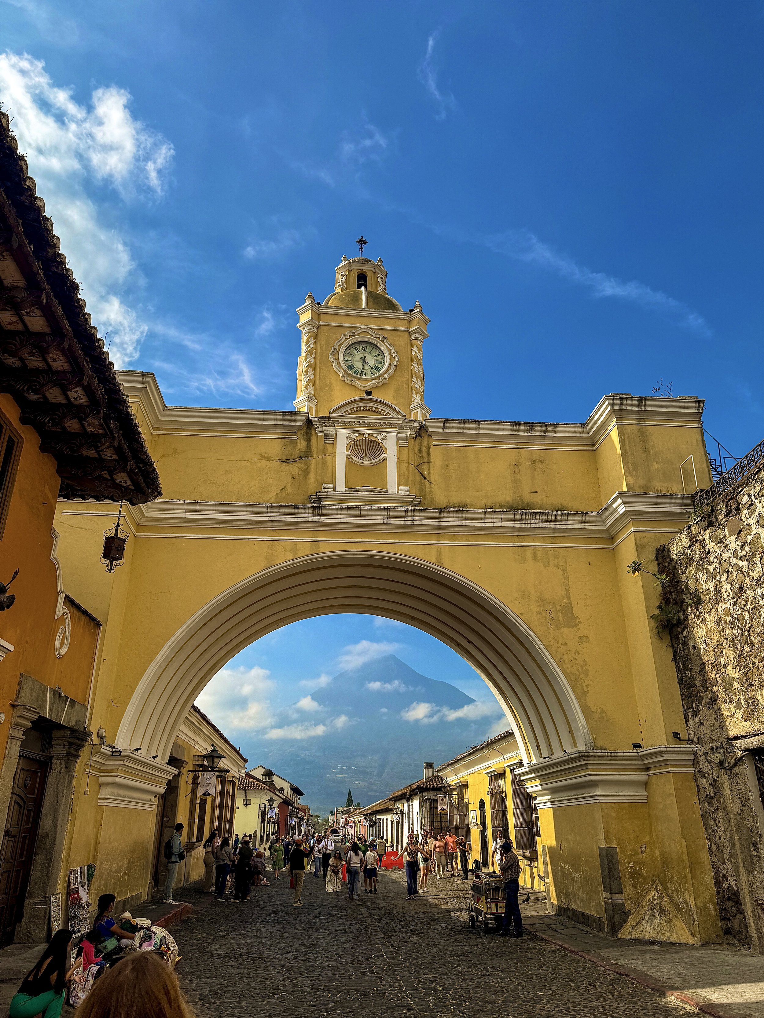 Street scene in Antigua Guatemala with a large arch framing a view of a distant volcano, yellow colonial buildings, and a bustling crowd of tourists and vendors.