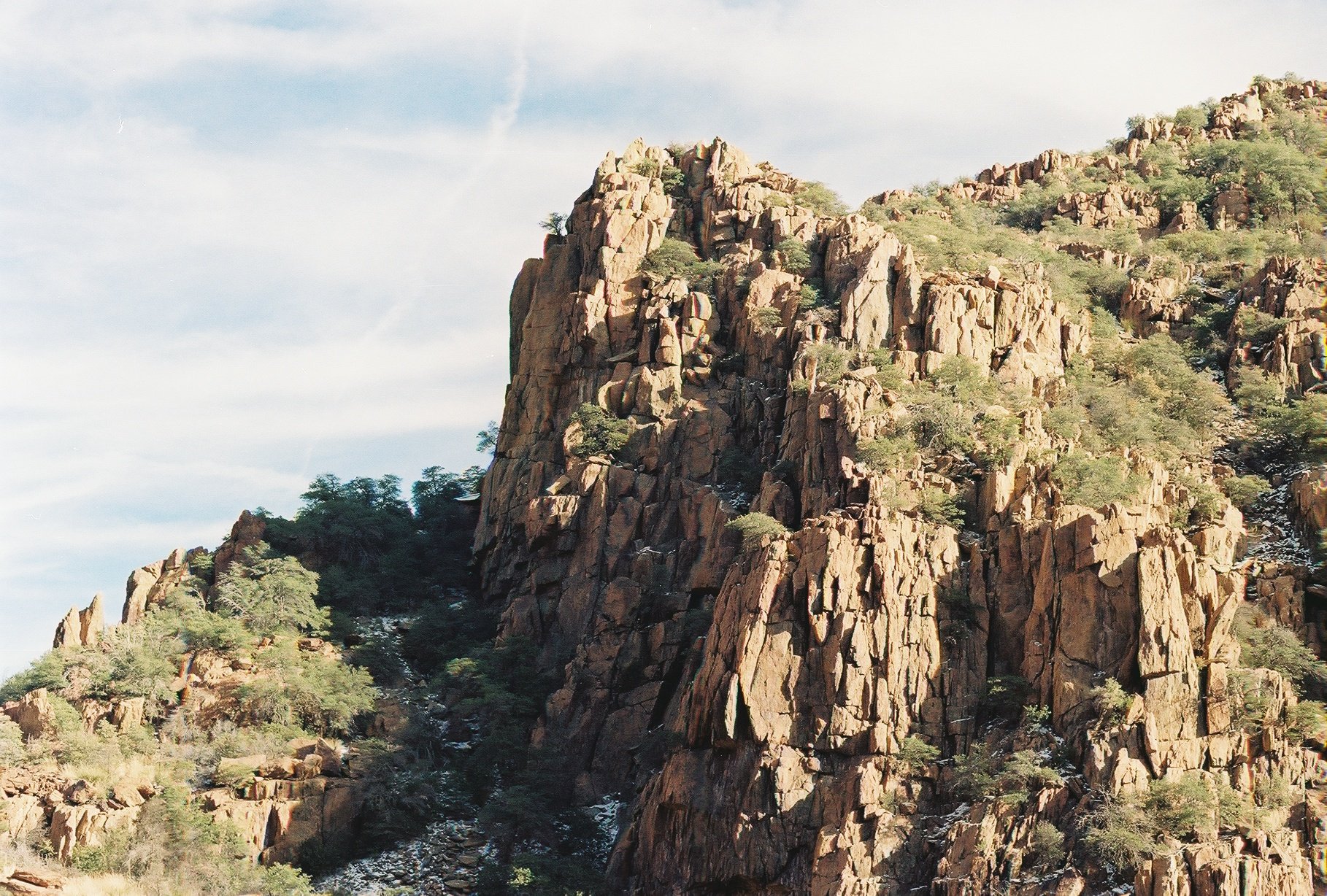A rugged mountain with steep cliffs covered in sparse green vegetation and small trees, under a partly cloudy sky.