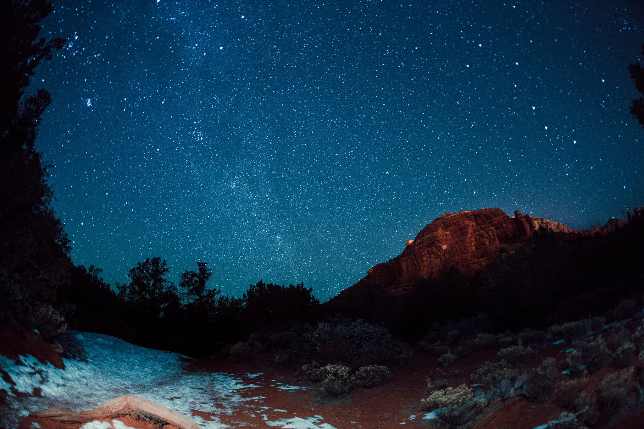 Night sky filled with stars above a rocky landscape with a river, cliffs, and trees in the foreground.