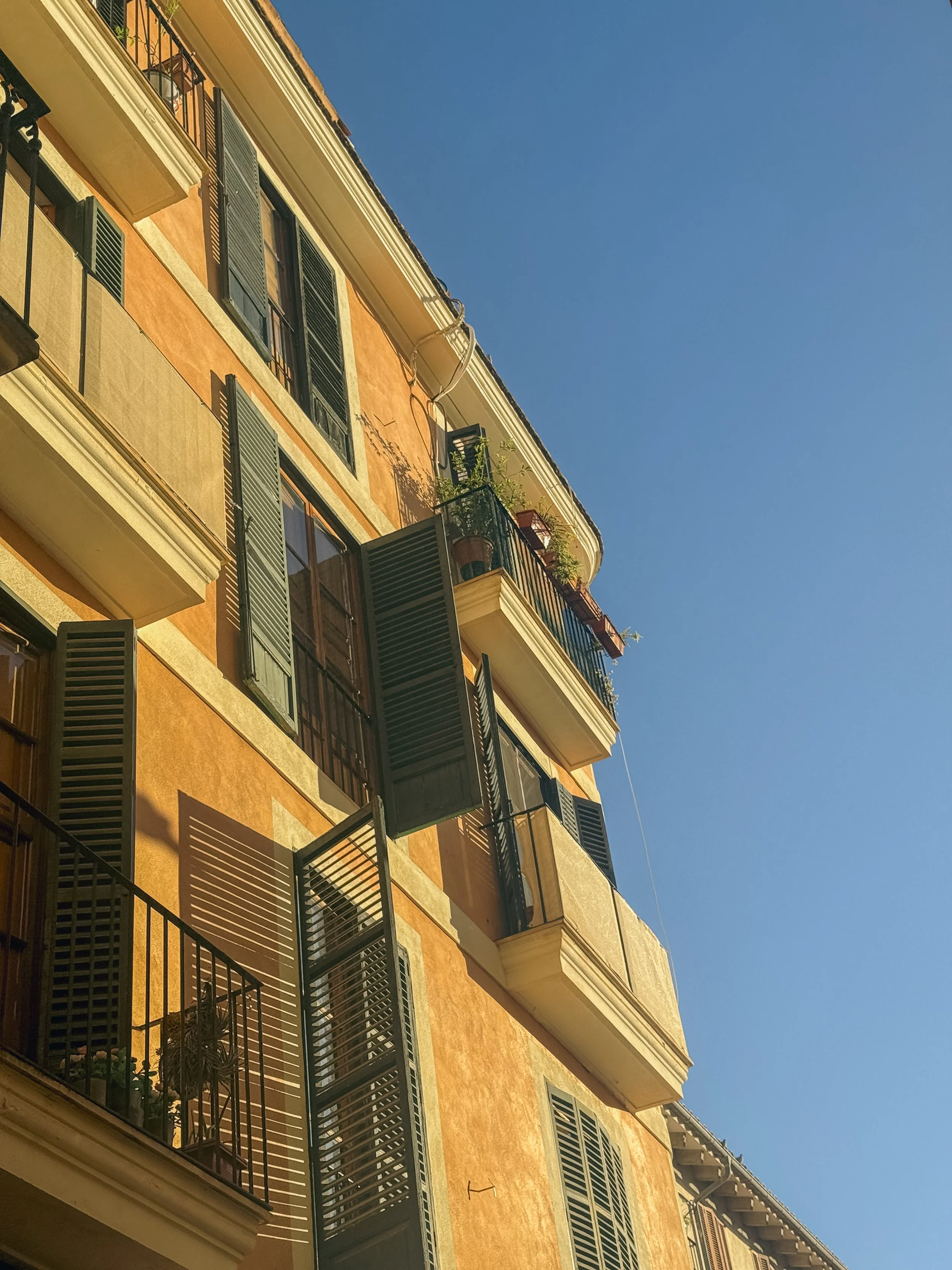 Multiple balconies with black railings and green shutters on a yellow-orange apartment building against a clear blue sky.
