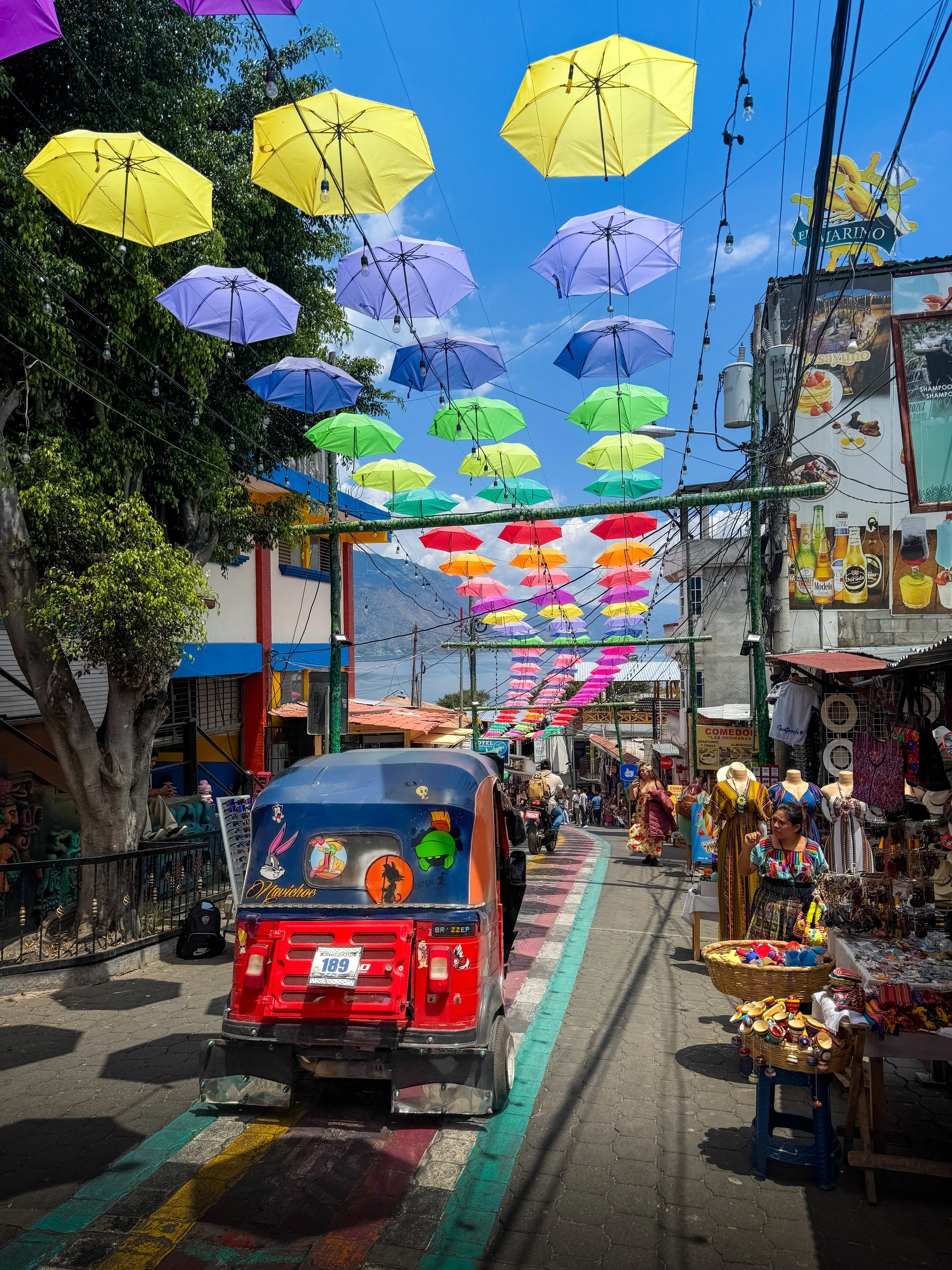 Street scene decorated with colorful umbrellas hanging overhead in a marketplace with shops and vendors selling clothes, hats, and souvenirs. A tuk-tuk vehicle is in the foreground, and people are walking along the street.