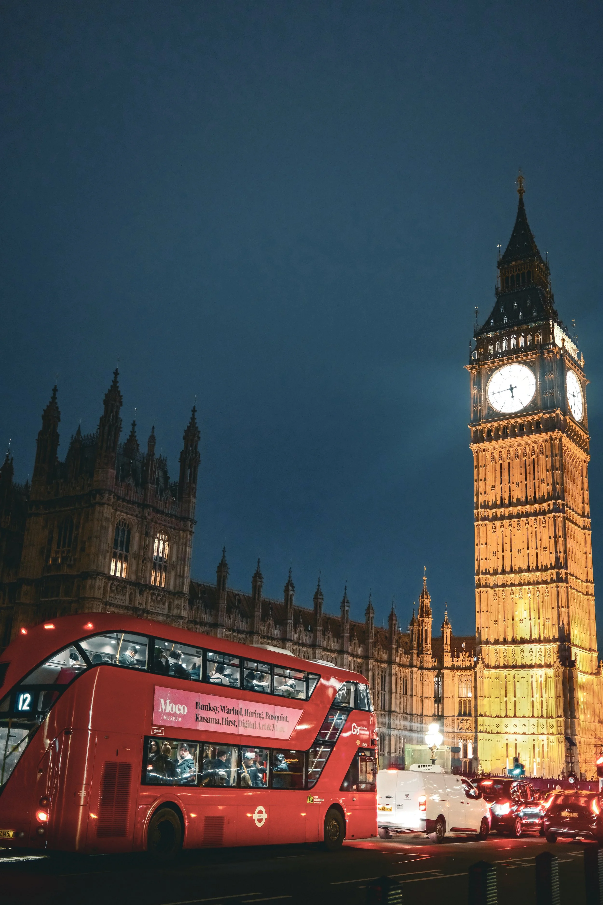Night view of Big Ben clock tower in London, illuminated against the dark sky, with a red double-decker bus and cars on the street in the foreground.