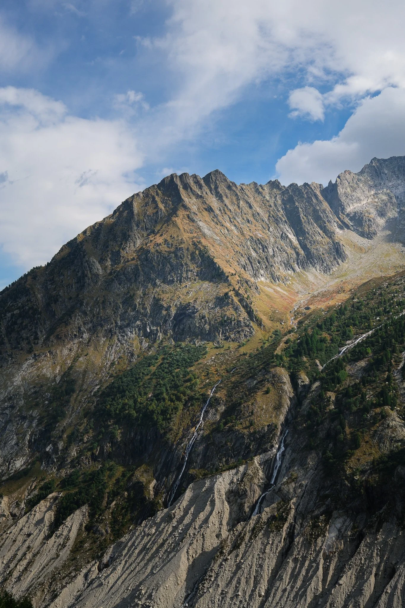 Landscape of tall mountain peaks with steep, rocky slopes, some small waterfalls flowing down, under a partly cloudy sky.