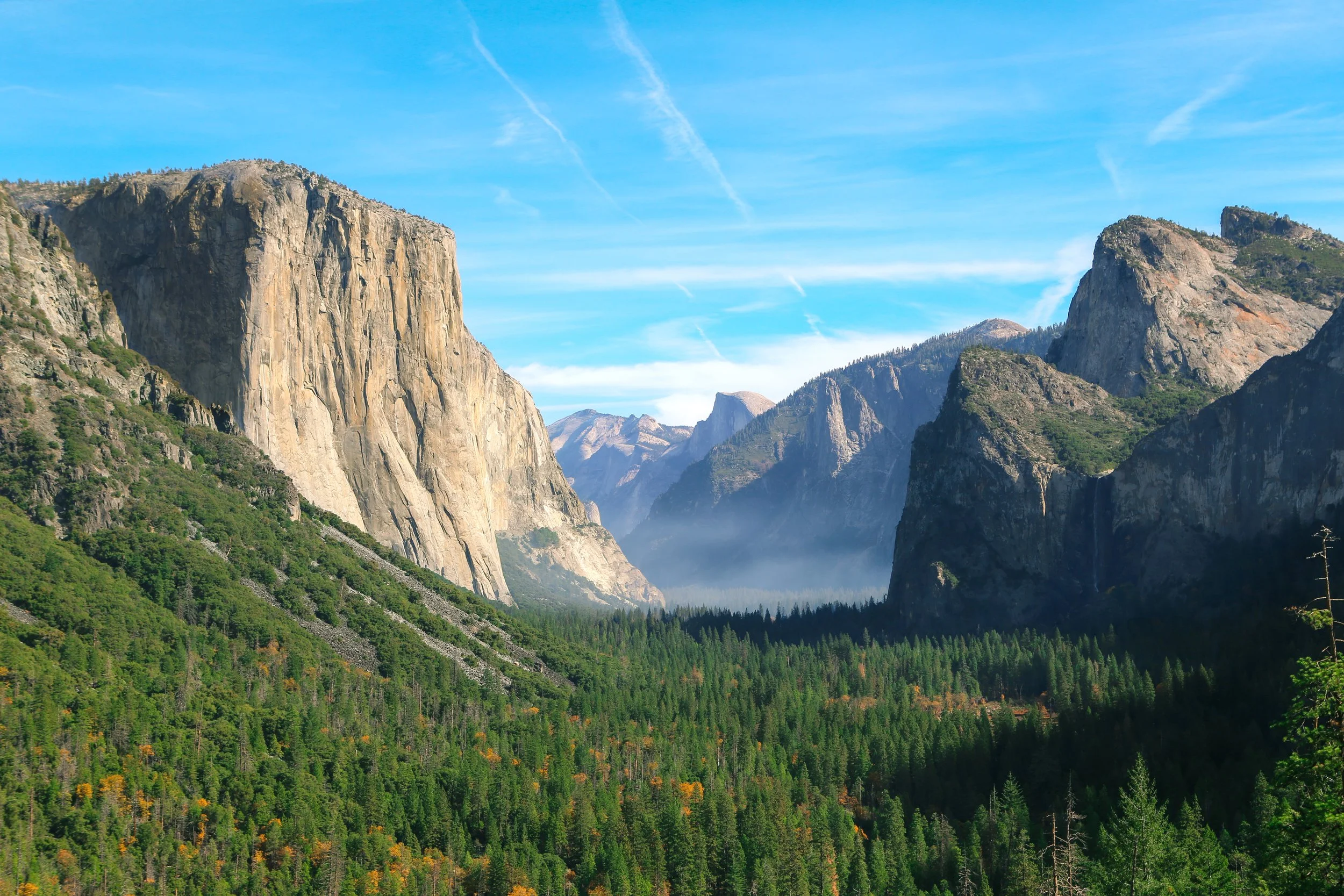 Scenic view of Yosemite Valley featuring tall granite cliffs, lush green forests, and a bright blue sky with a few wispy clouds.