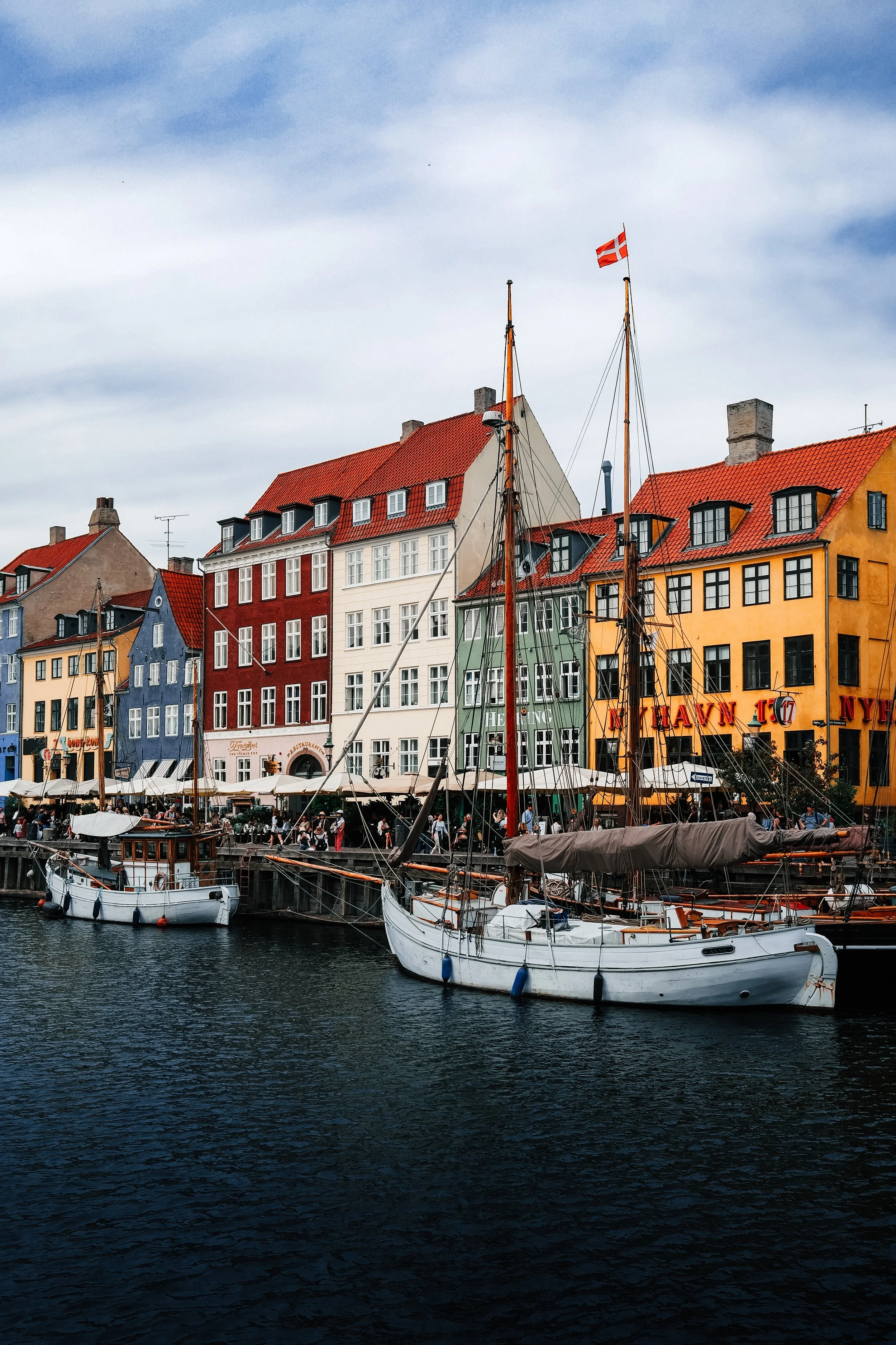 Colorful buildings with red, yellow, blue, and green facades along a waterfront with sailboats moored in the water and a Danish flag flying on a boat. Overcast sky in the background.