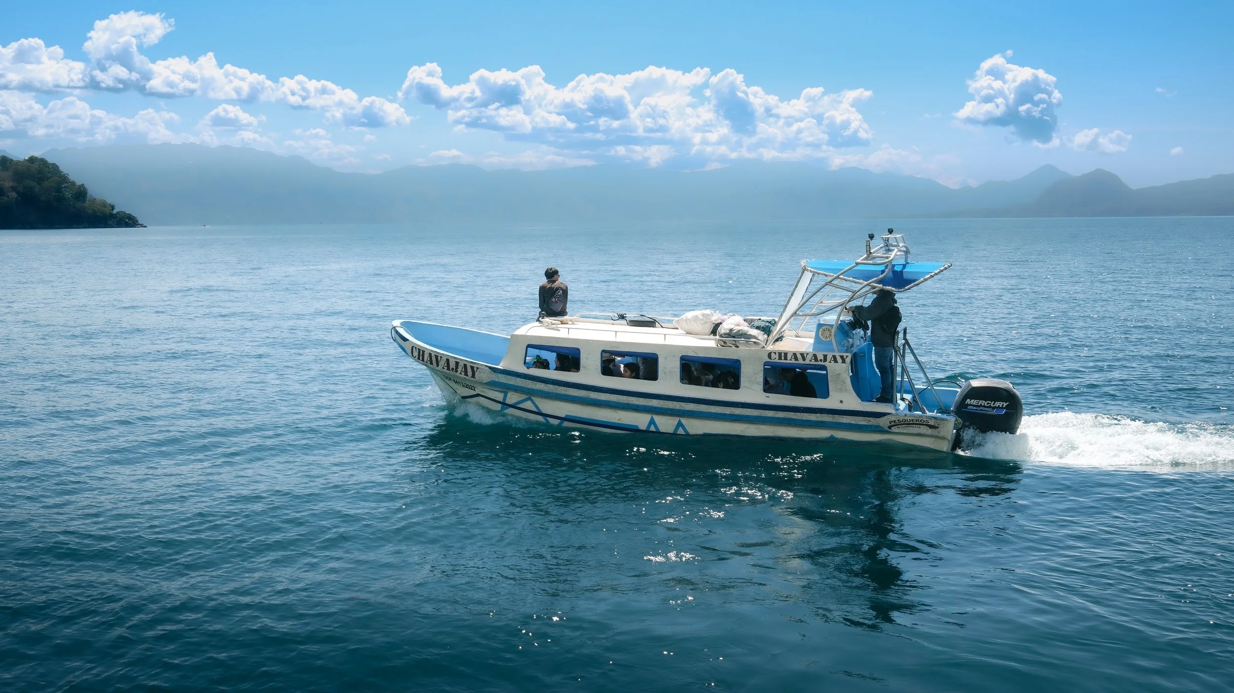 A boat named 'Chavajay' with people on board sailing across a large body of water under a partly cloudy sky, with mountains in the background.