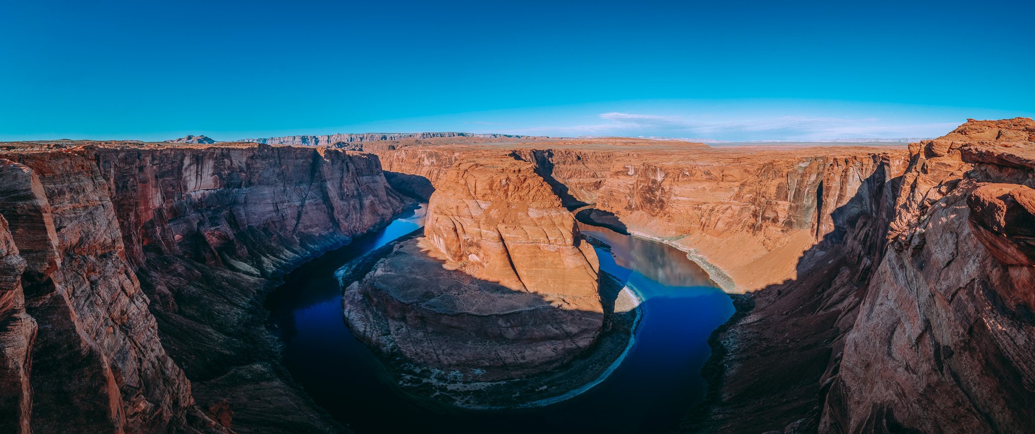 A wide view of the Grand Canyon with the Colorado River winding through the canyon, steep red rock cliffs on either side, and a clear blue sky above.