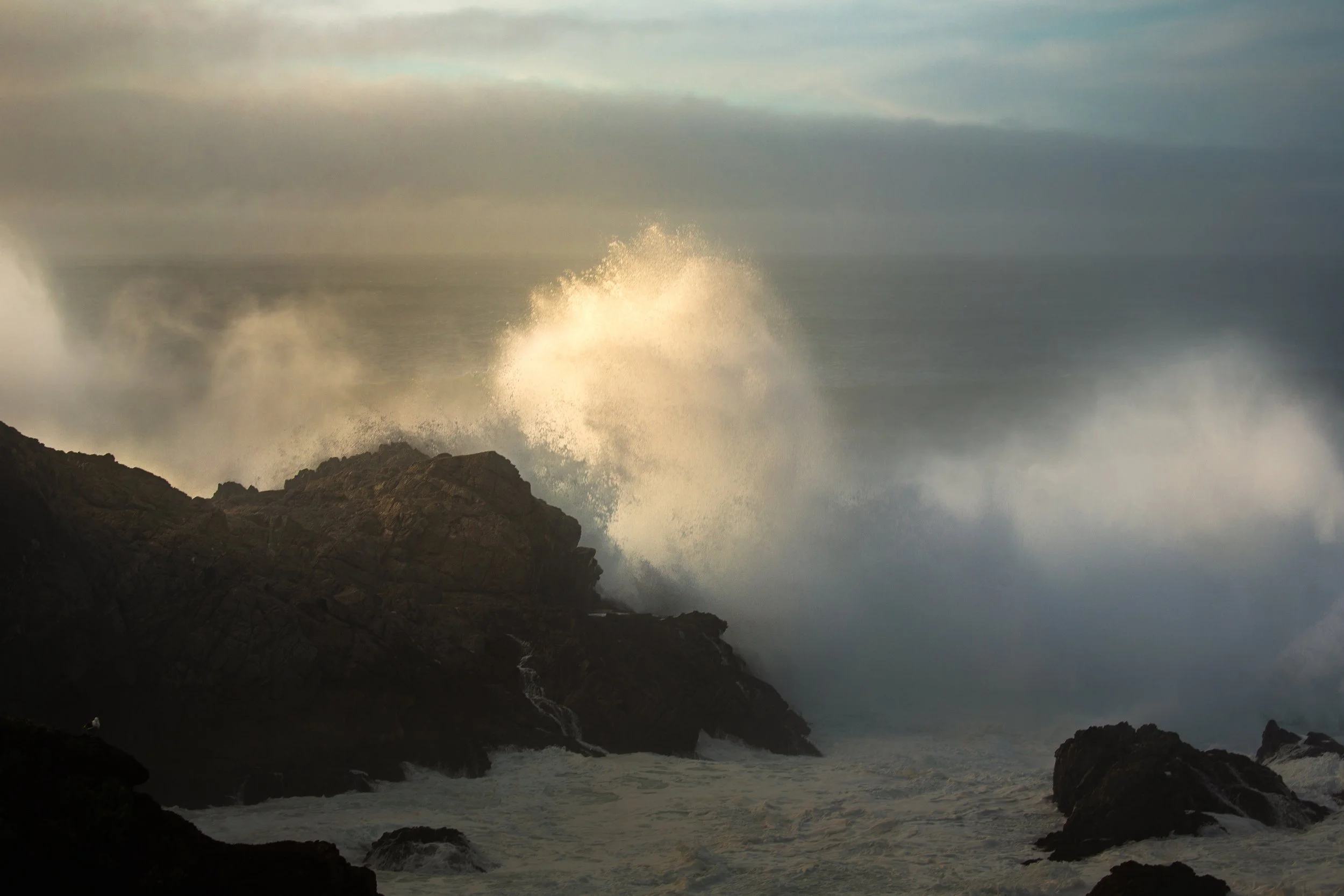 Powerful ocean waves crashing against rugged rocks during a cloudy day, with mist and spray rising into the air.