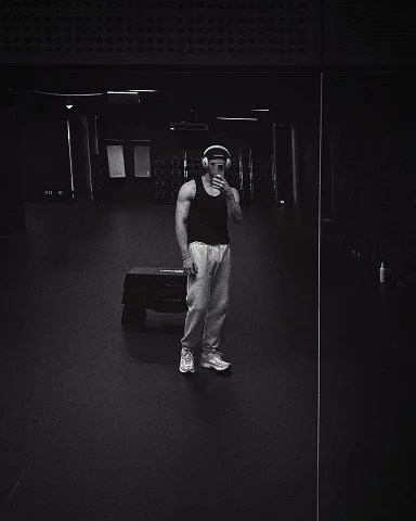 Person taking a mirror selfie in a gym, wearing a black sleeveless shirt, sweatpants, and sneakers, with gym equipment in the background.