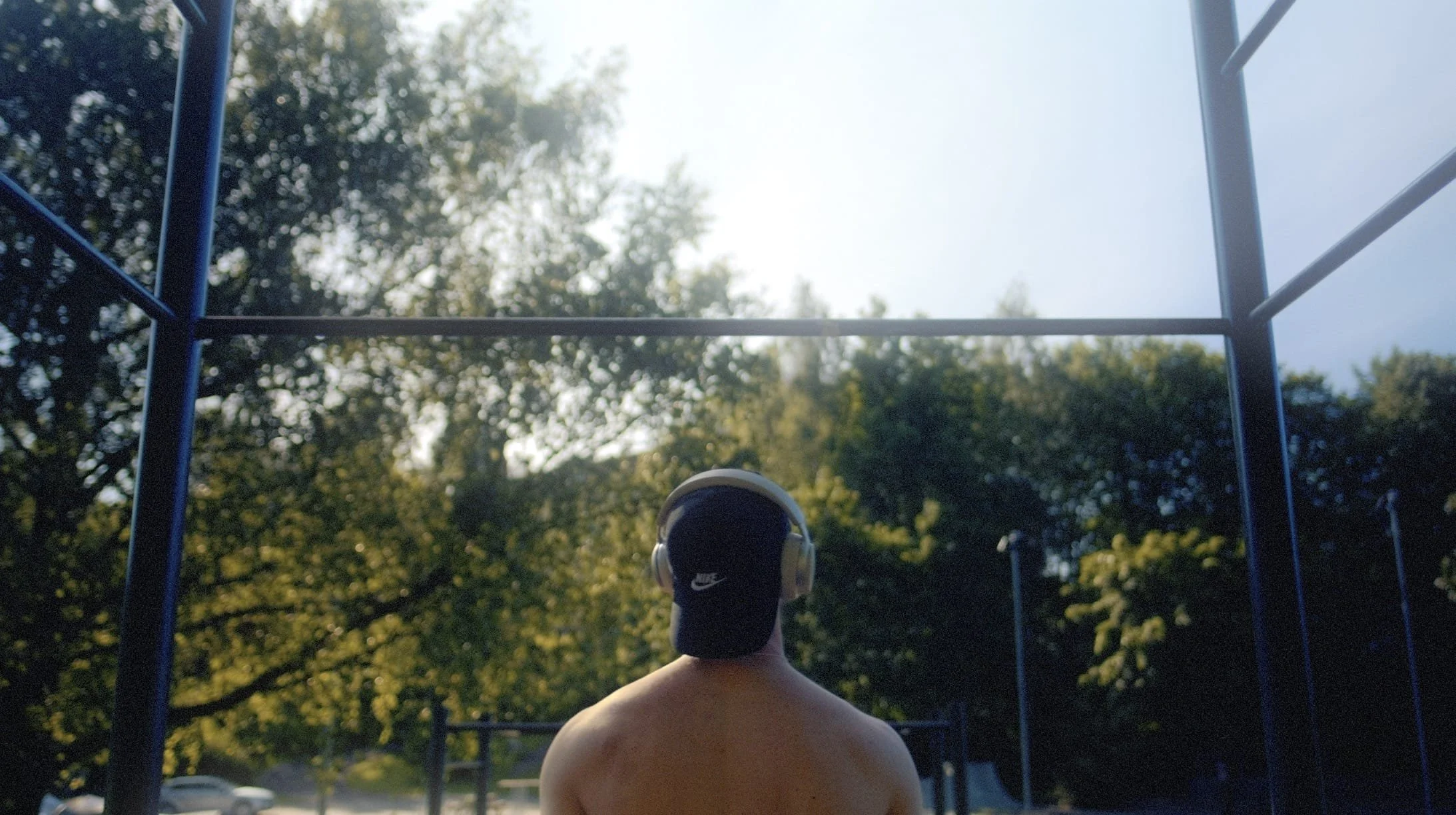 A person with headphones standing outdoors facing a playground structure with trees and sky in the background.
