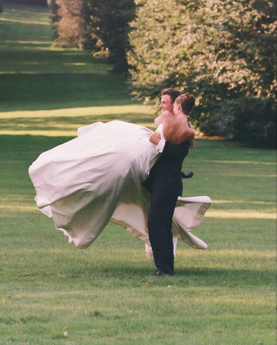 A bride in a white wedding gown being lifted by a groom in a black tuxedo on a grassy field with trees in the background.