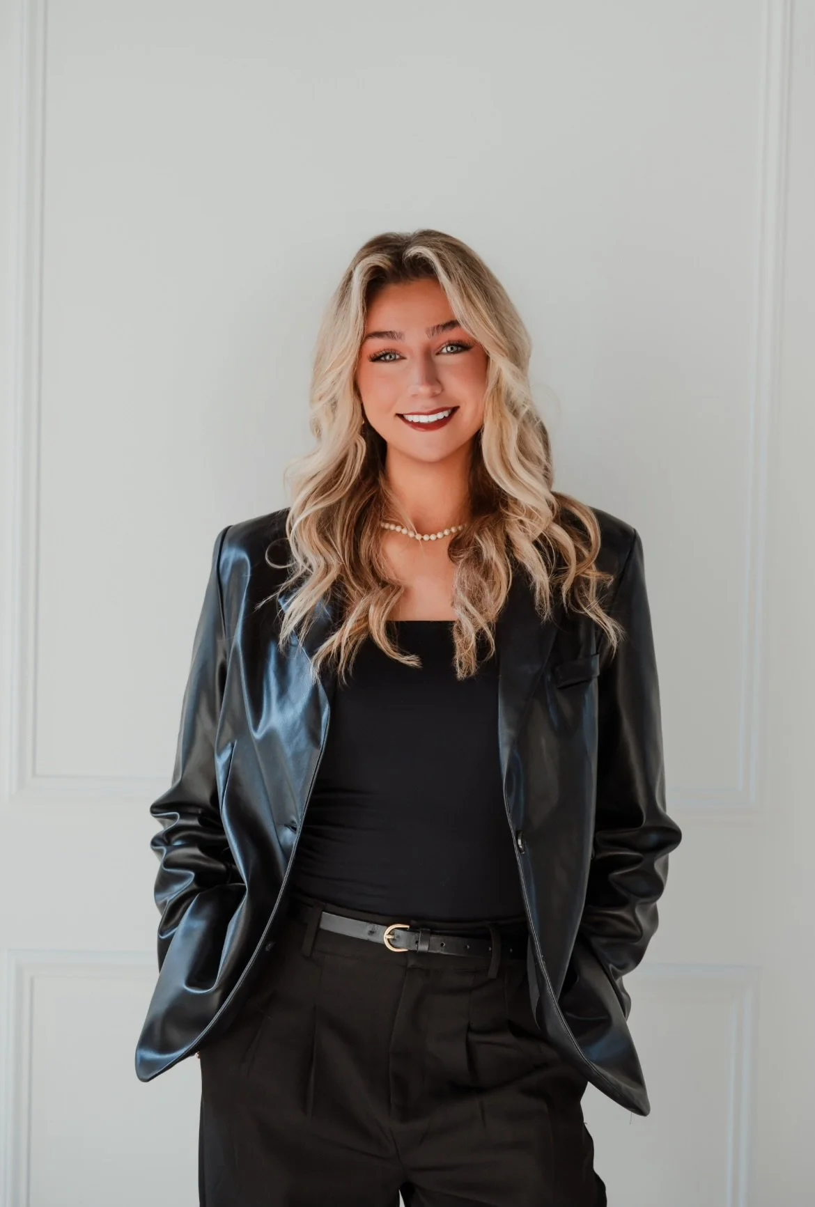 A woman with wavy blonde hair, wearing a black leather jacket, black top, and black trousers, standing in front of a plain white wall with subtle paneling, smiling.