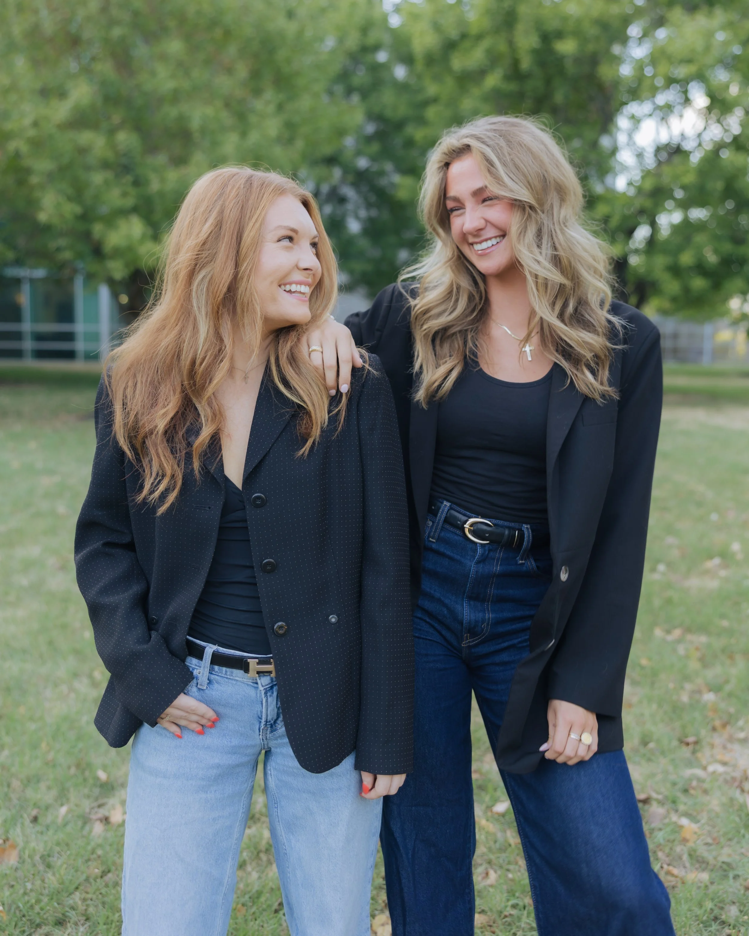 Two women smiling, standing outdoors on grass with green trees in background, one with red hair and light blue jeans, the other with blond hair and dark blue jeans, wearing dark blazers over black tops.