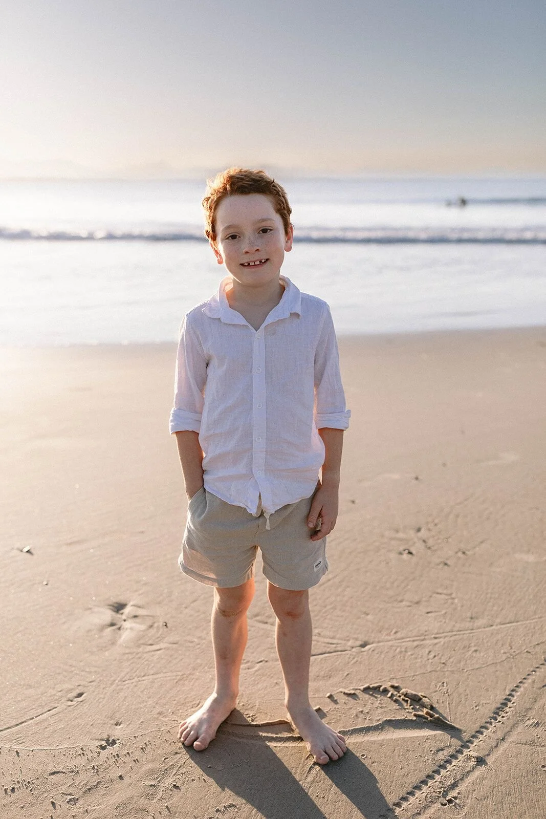 Boy in a white shirt and khaki shorts standing barefoot on the sand at sunset